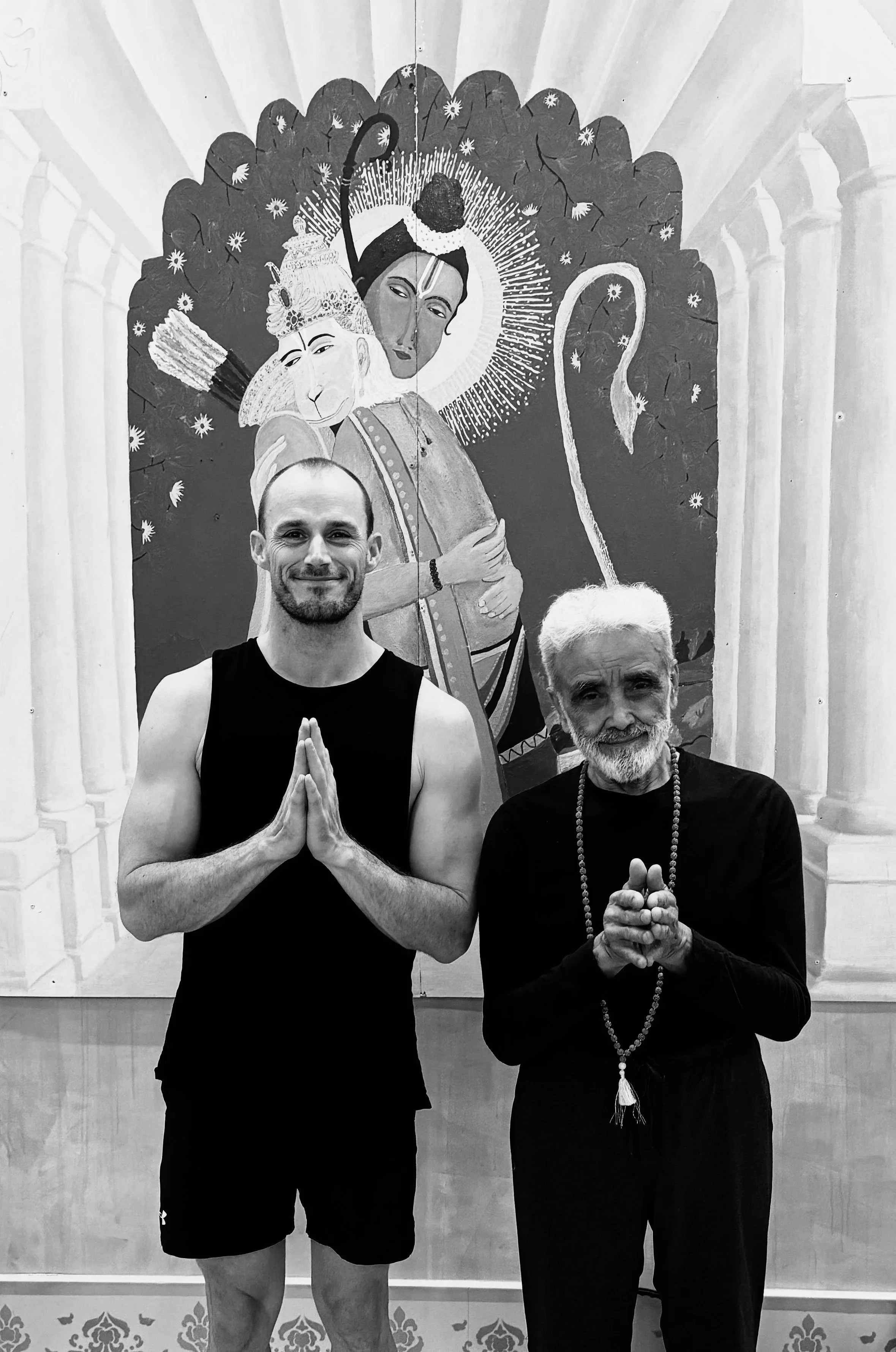 Two men stand in front of a religious or spiritual painting, with their hands in prayer-like gestures. The man on the left is young, bald, and wearing a sleeveless shirt and shorts. The man on the right is older, with white hair and a beard, dressed in black with a beaded necklace.