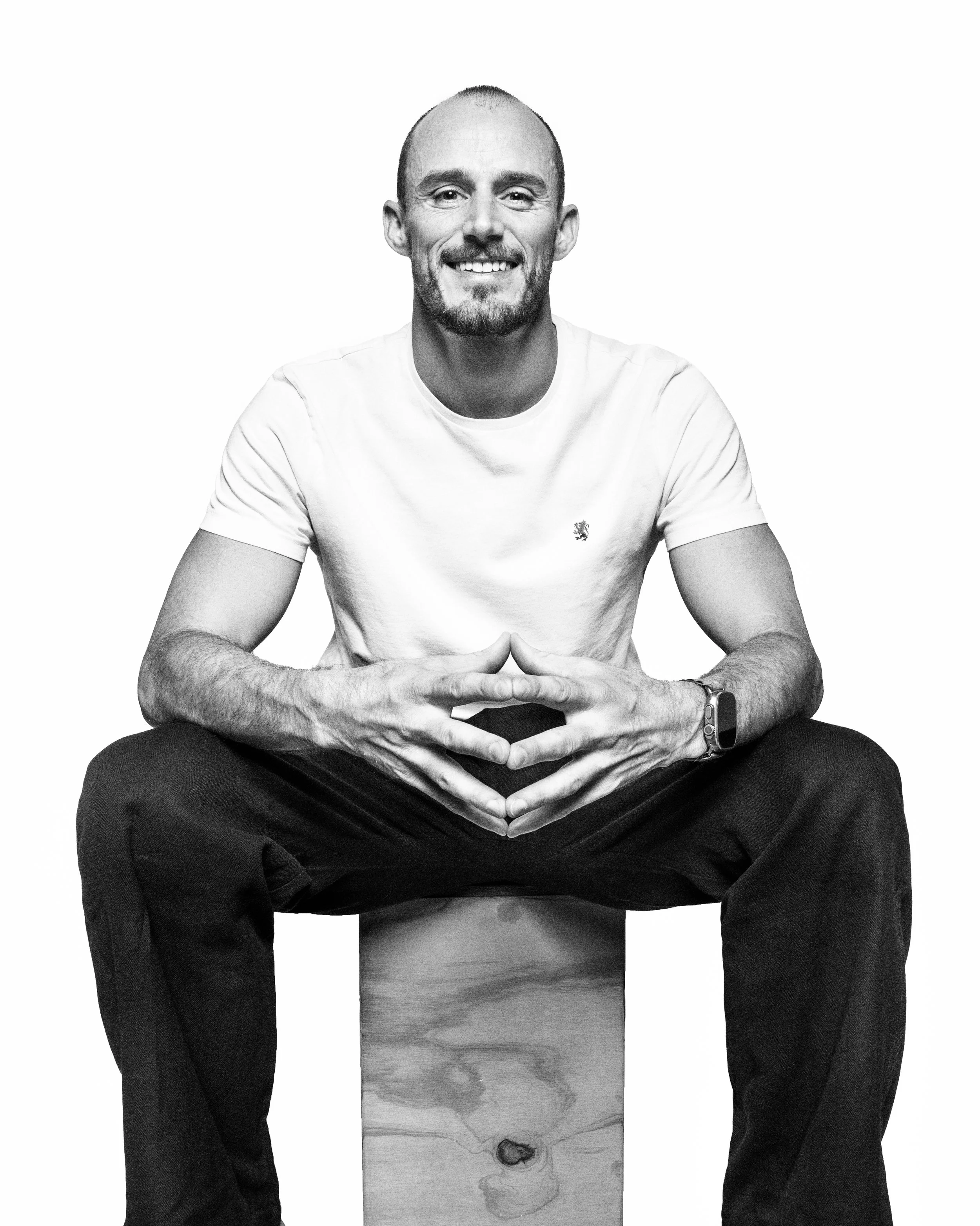 A man with a beard and shaved head, smiling, sitting cross-legged on a wooden block, wearing a white t-shirt and dark pants, against a plain white background.