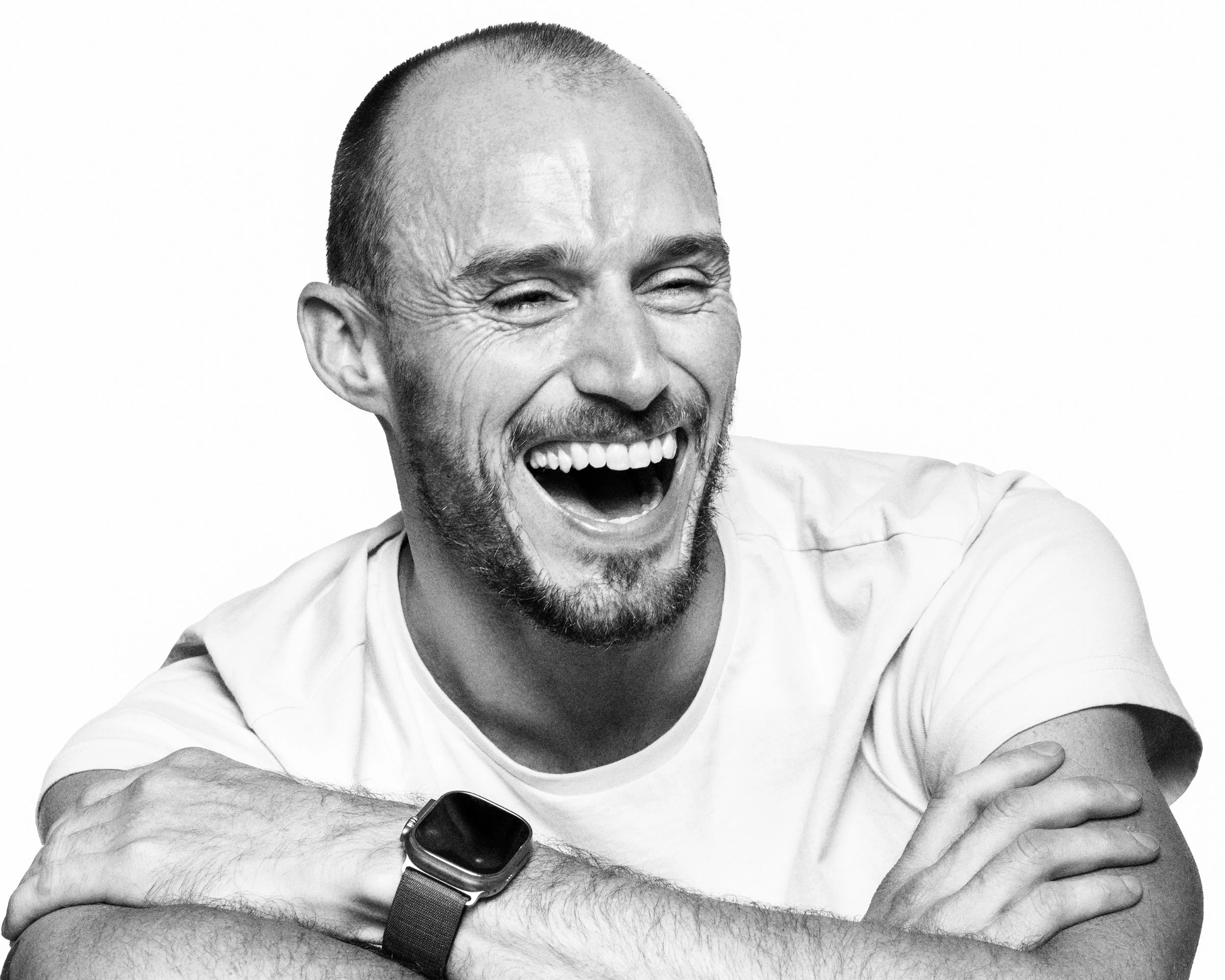 A man with a beard and short hair laughing with arms crossed, wearing a smartwatch and a white t-shirt.