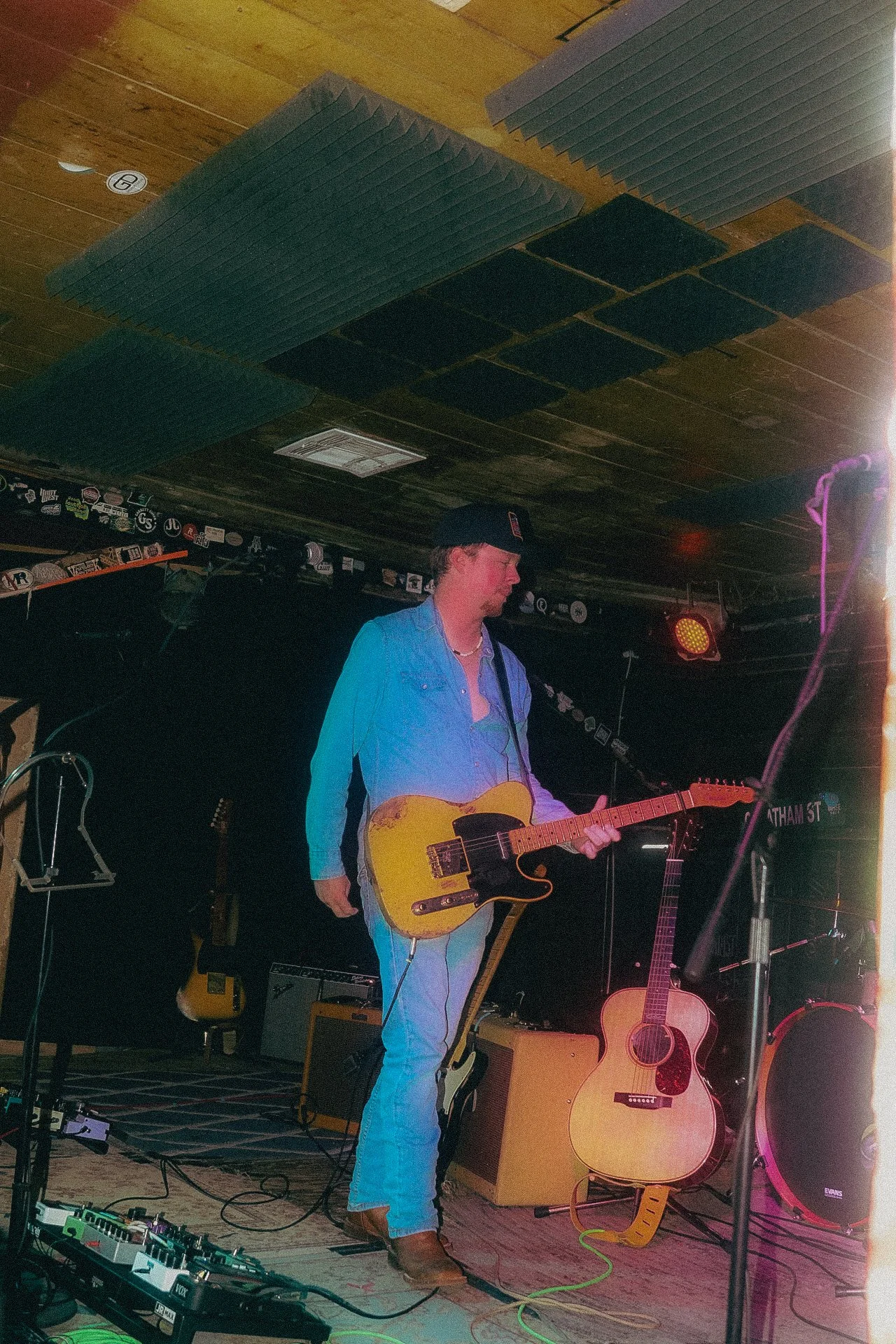 A male musician wearing a black cap, light blue denim shirt, and jeans playing an electric guitar on stage in a venue with musical equipment and acoustic guitars nearby.