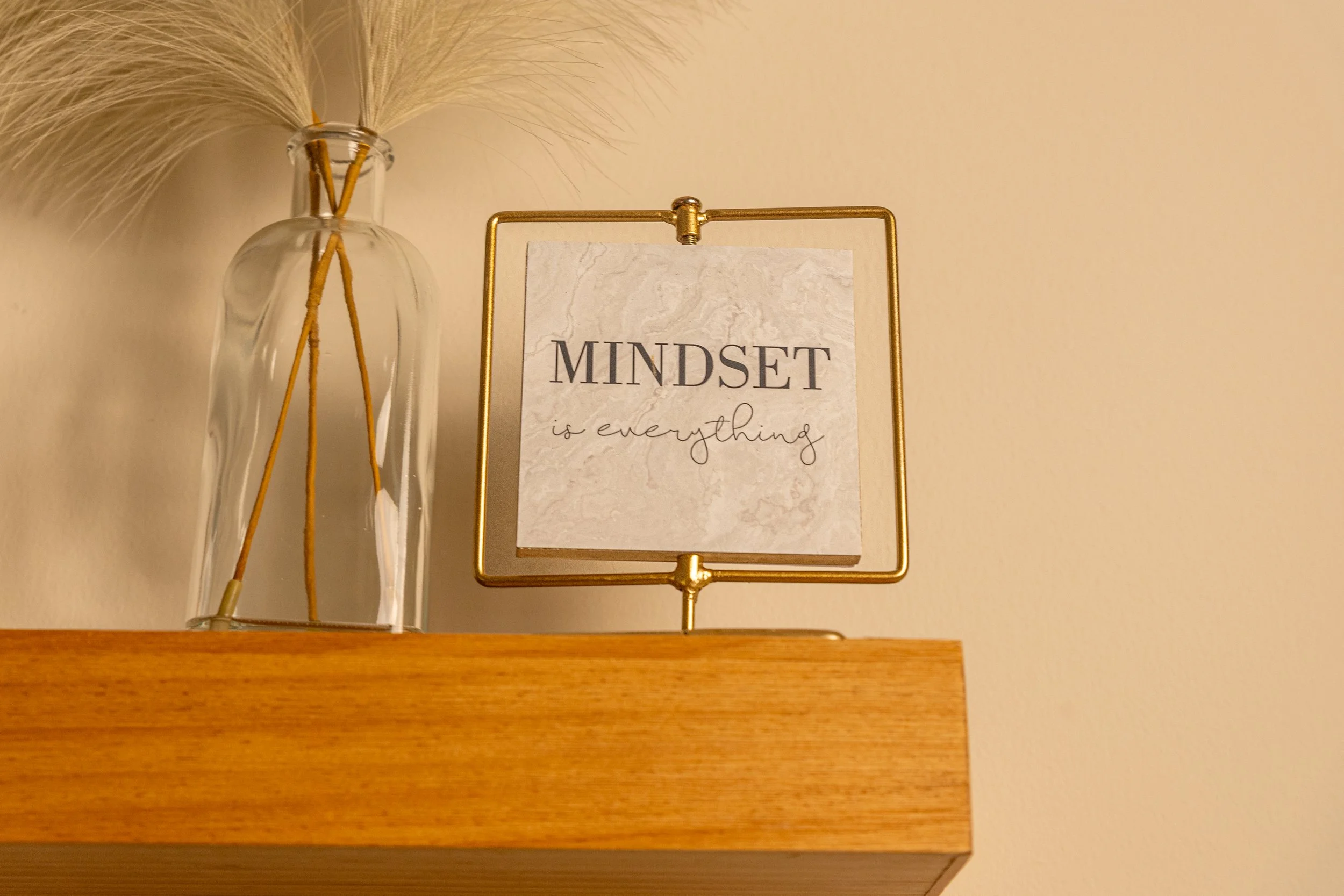 Decorative shelf with a clear glass vase holding pampas grass and a framed quote that says "Mindset is everything" on a beige wall.