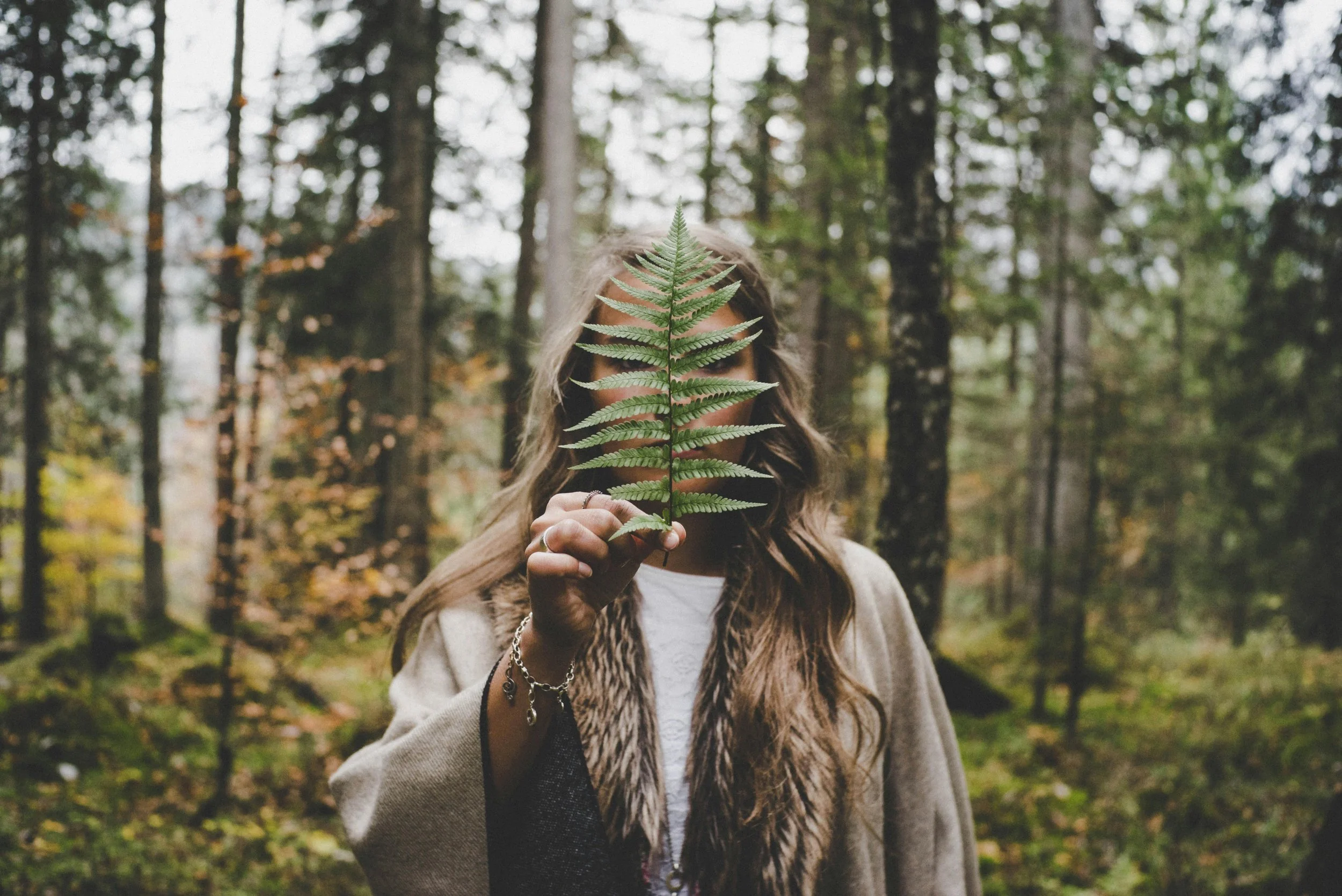 A person holding a fern leaf in front of their face in a forest with tall trees.
