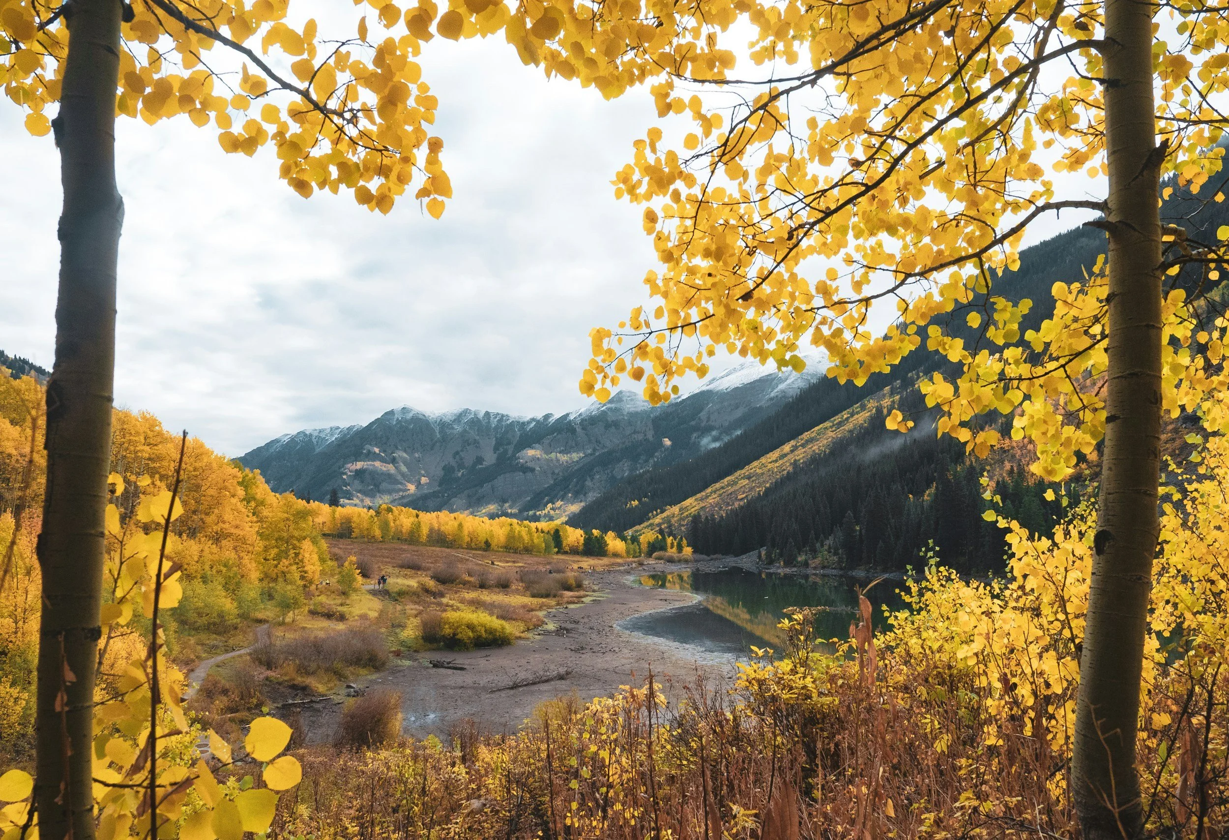 A scenic landscape of a mountain valley with a river, surrounded by trees with yellow autumn leaves and snow-capped mountains in the background.