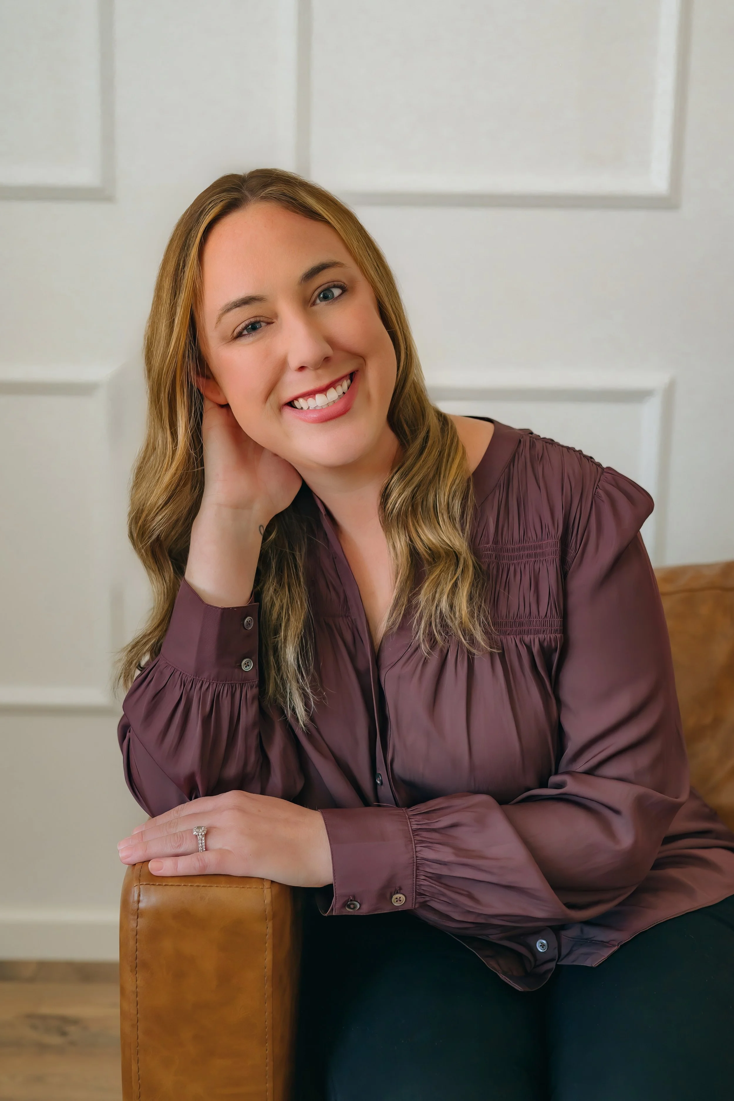 A woman with wavy light brown hair, wearing a long-sleeve purple blouse, smiling with her head tilted, sitting on a leather chair in front of a white wall with decorative panels.
