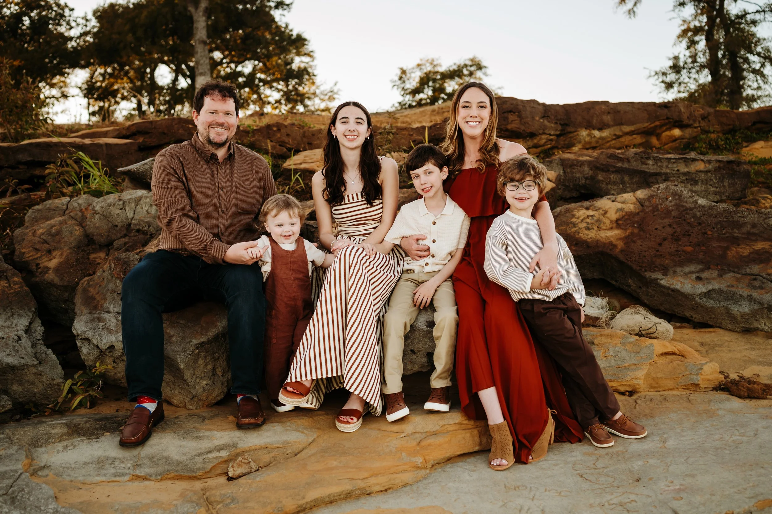 A family of seven sitting on rocks outdoors, smiling at the camera during sunset, in a natural setting with trees in the background.