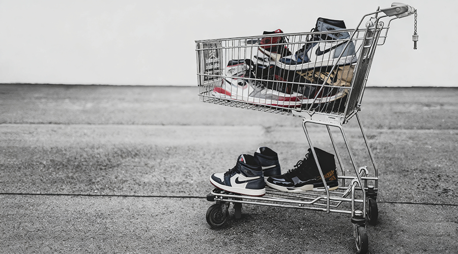 A shopping cart filled with various sneakers, with some sneakers placed on the ground in front of the cart.