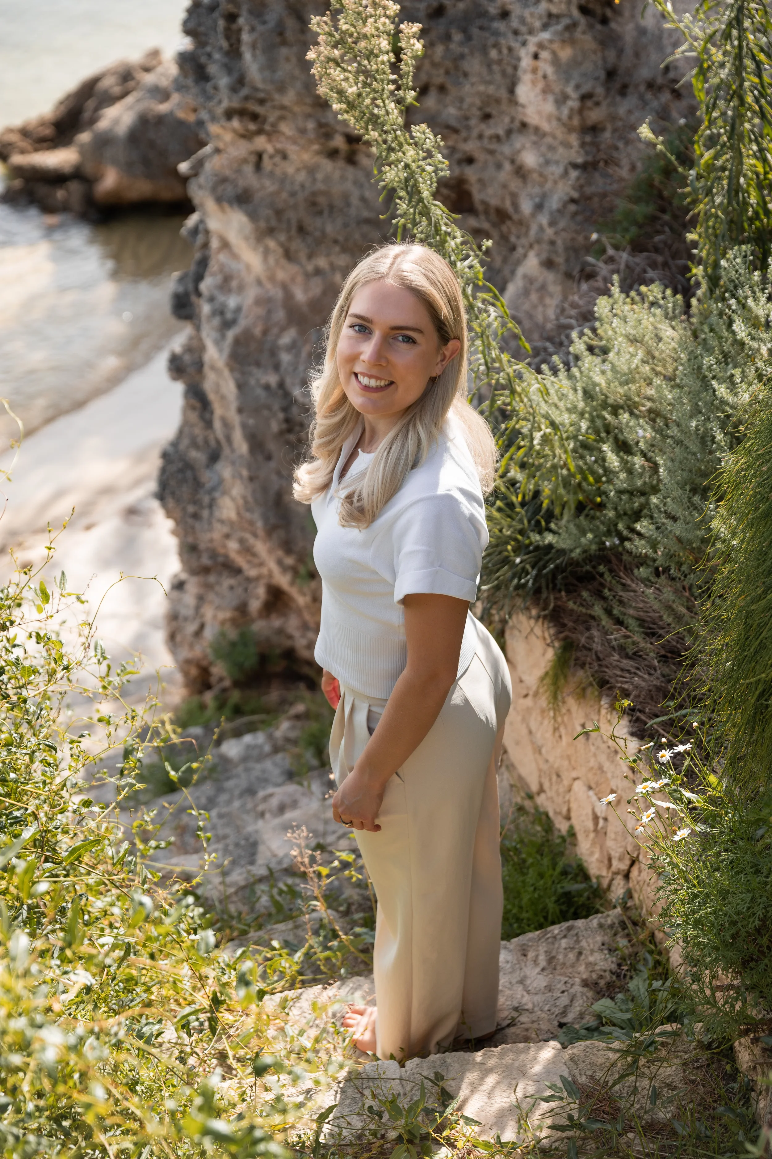A woman with blonde hair smiling and looking at the camera standing on rocky terrain outdoors, surrounded by green plants, near a body of water.