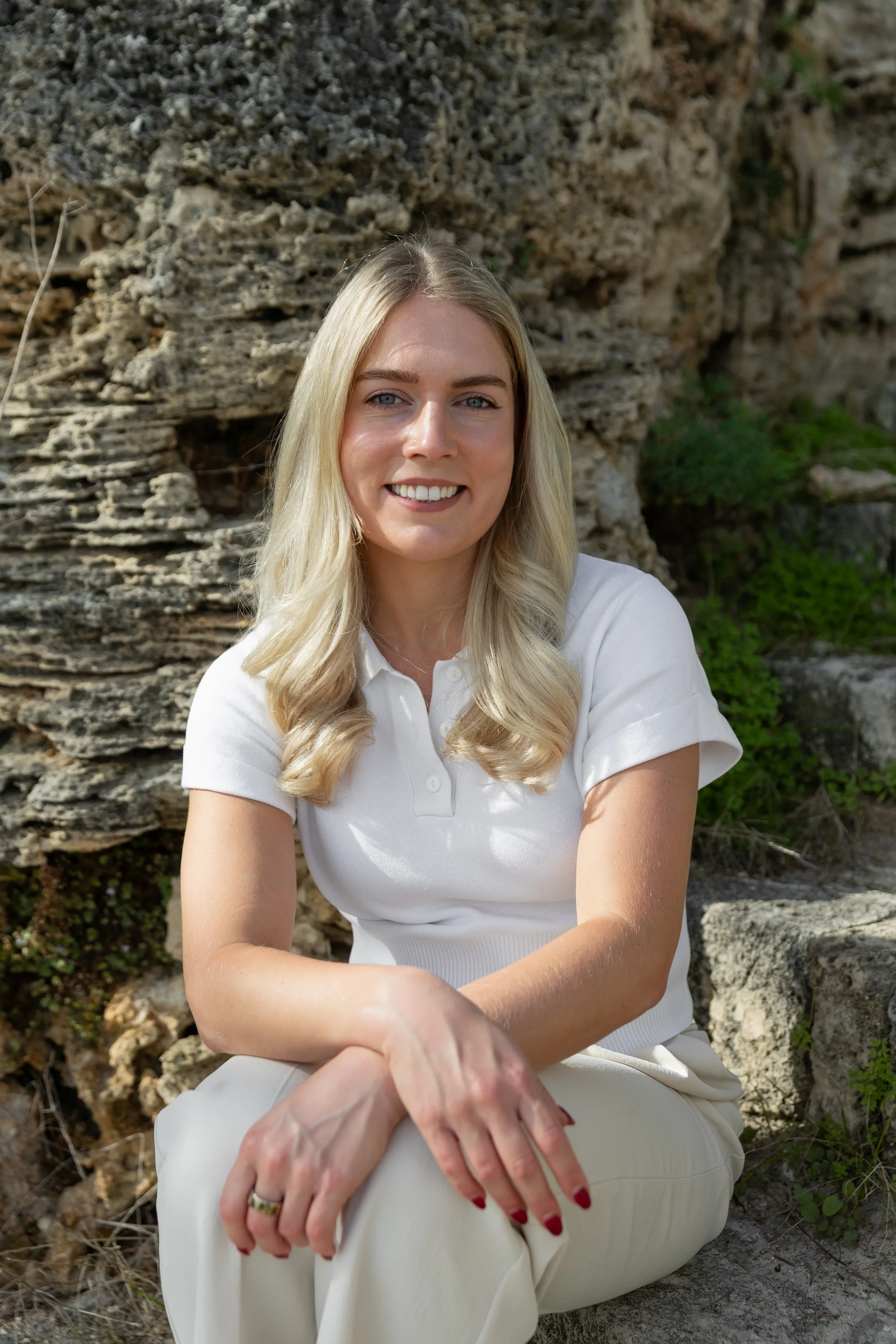 A young woman with long blonde hair, wearing a white shirt and cream pants, sitting outdoors against a rocky background with green foliage, smiling at the camera.