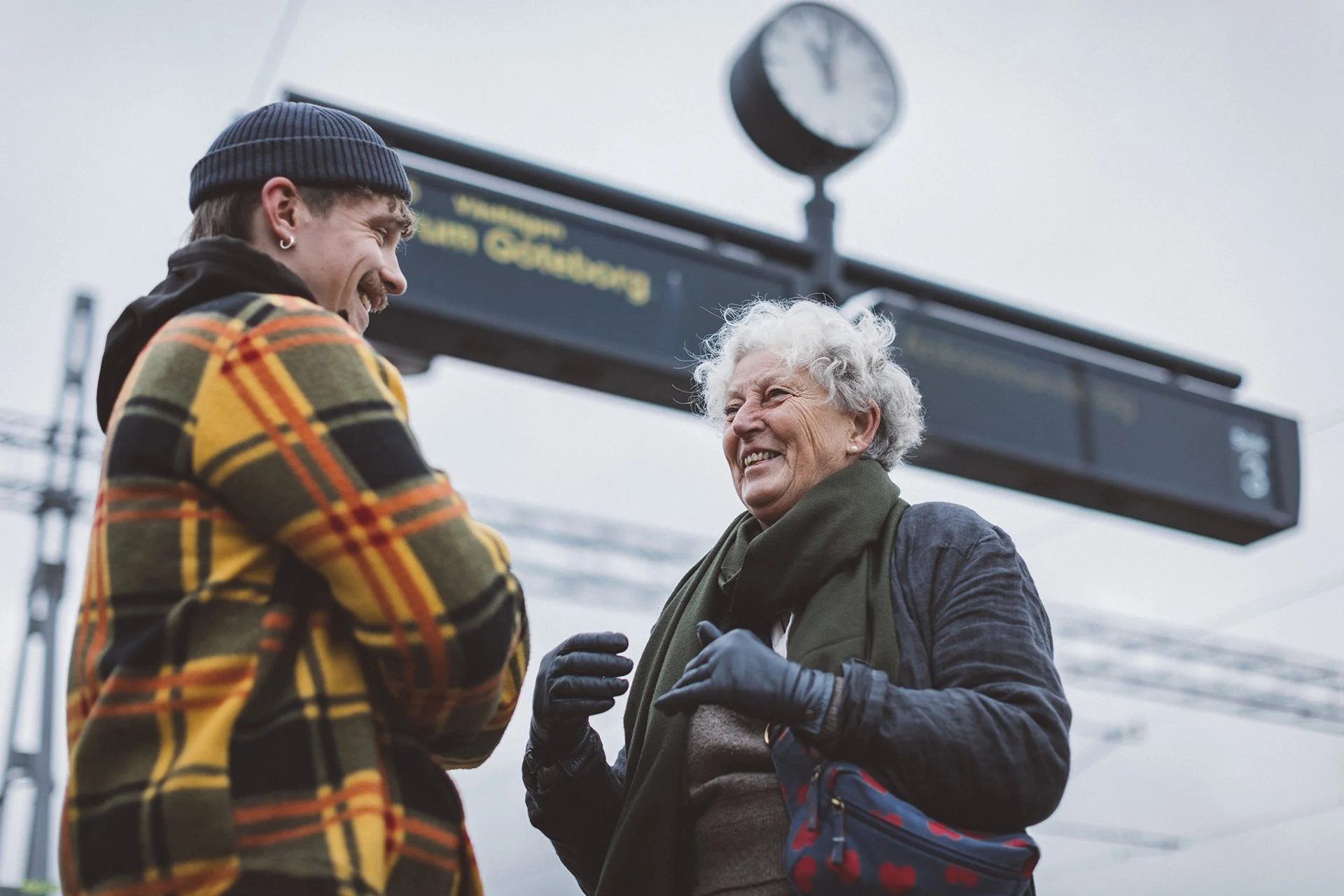 En ung man och en äldre kvinna står och pratar på en tågstation med ett tågskyltskylt i bakgrunden.