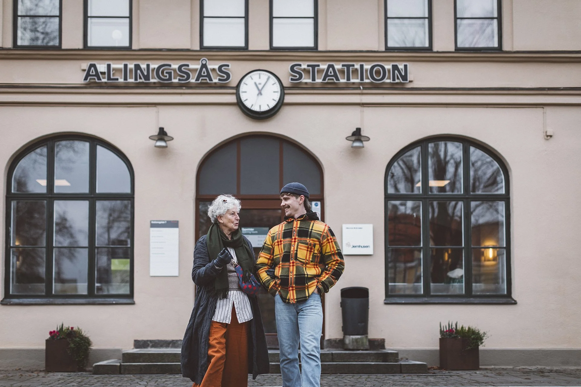 En äldre kvinna och en ung man som står utanför Alingsås stations byggnad och pratar.