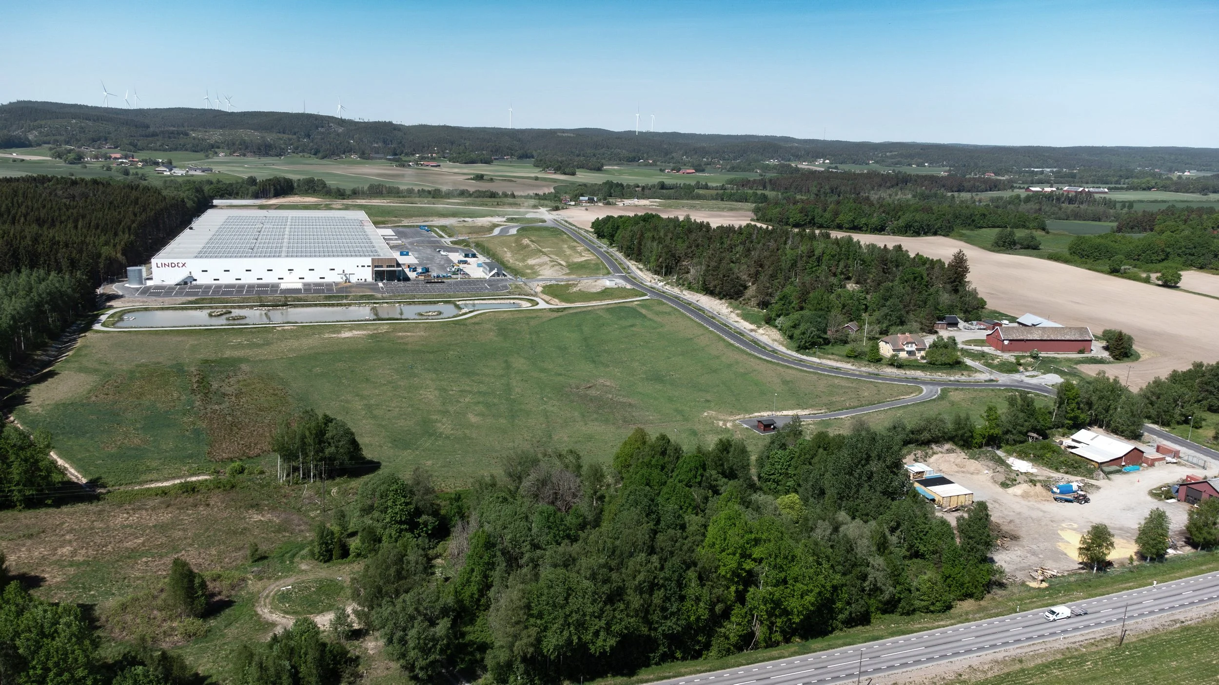 Vy av ett landskap med ett stort vitt lagerhyreshus, omgiven av gröna fält, skog och landsbygd, samt en väg och några små byggnader.