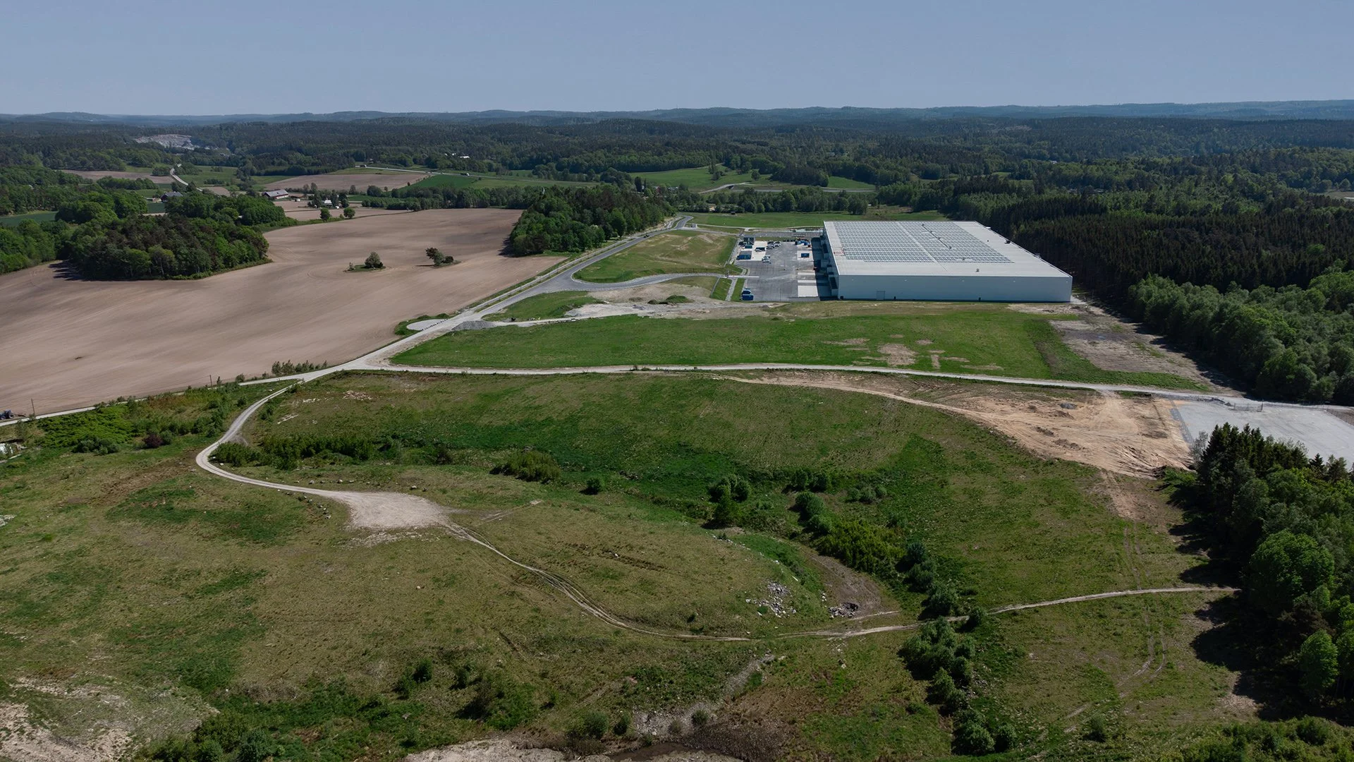 Vy av ett landskap med ett stort vitt lagerhyreshus, omgiven av gröna fält, skog och landsbygd, samt en väg och några små byggnader.