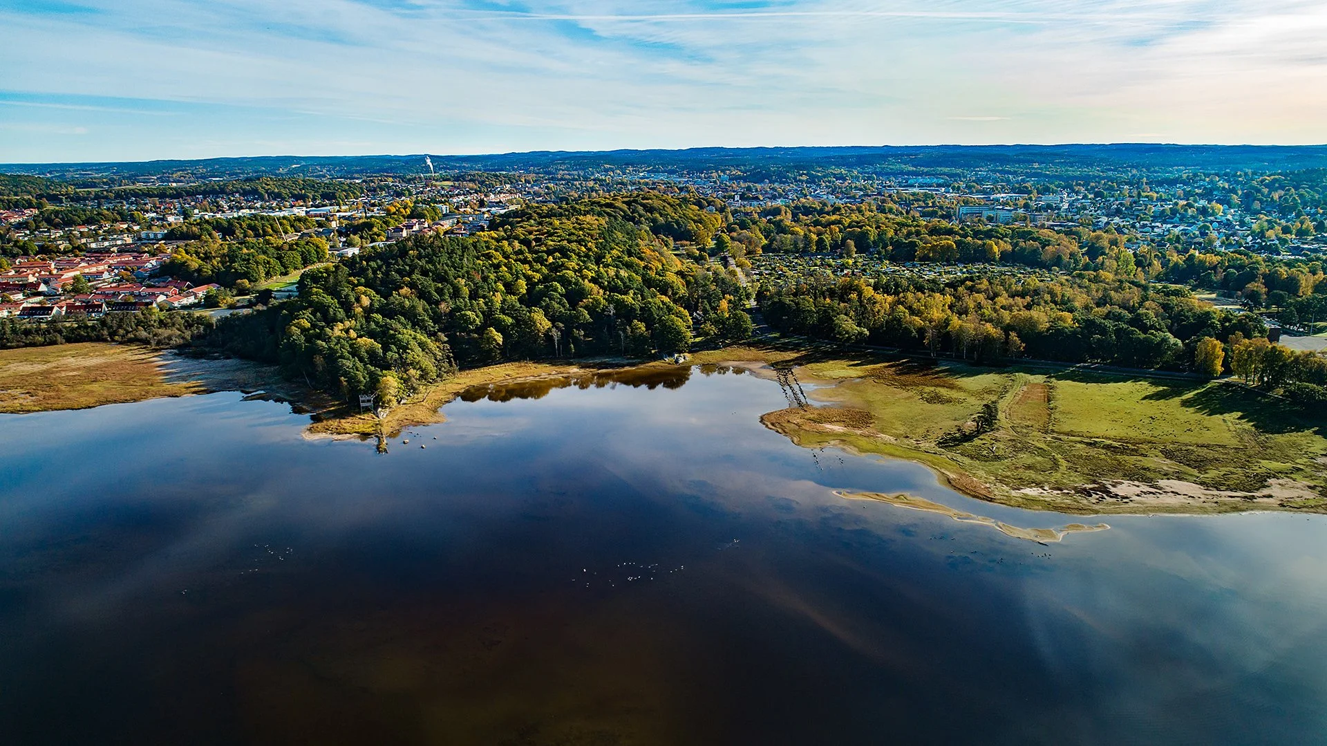 Flygbild av en sjö med omgivande skog och stadsutveckling i bakgrunden.