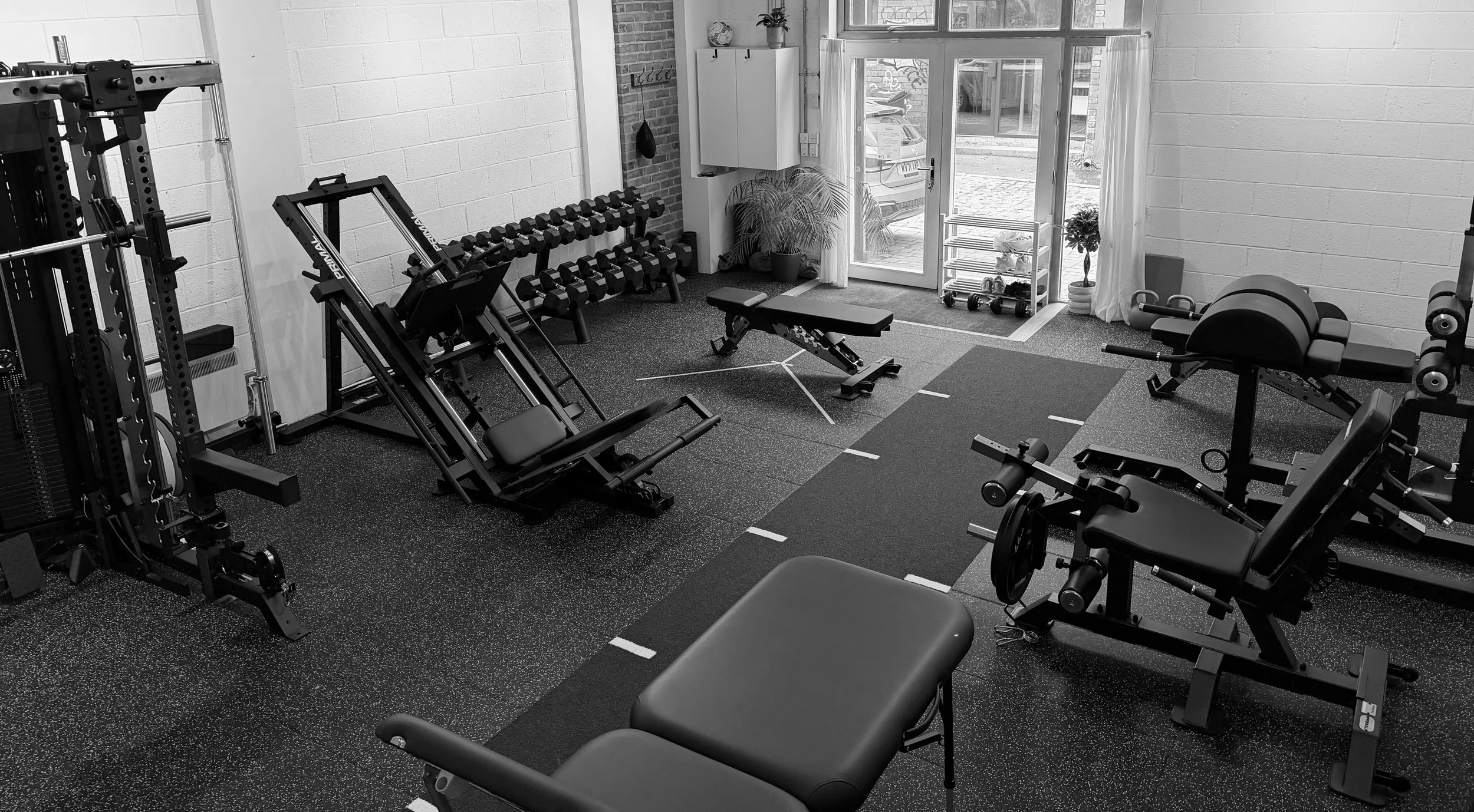 A clinic gym space with various workout equipment, including weight benches, dumbbells, a leg press machine, a squat rack cable machine, GHD, and knee extension/hamstring curl machine, and sled track.