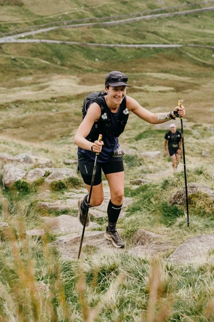 Woman hiking on rocky trail in grassy mountainous terrain, smiling, wearing a cap, sunglasses, hydration vest, shorts, and using trekking poles.