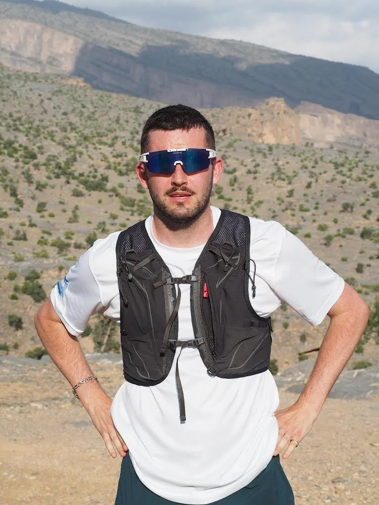 Man in sunglasses and a white t-shirt with a black hydration vest outdoors with mountains and sparse vegetation in the background.