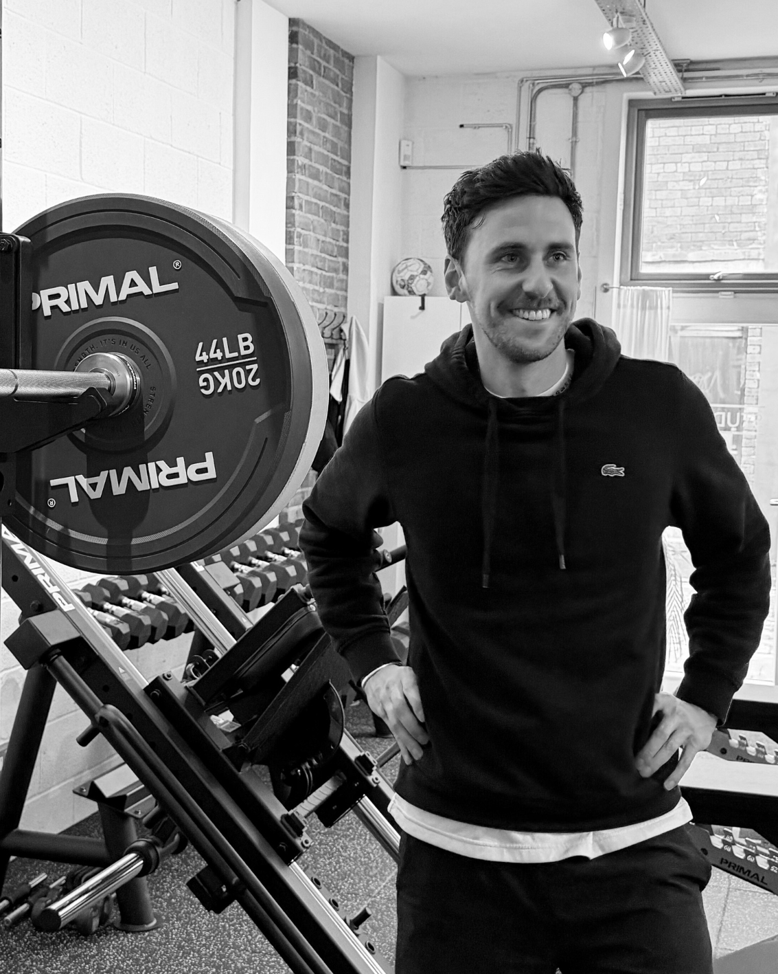 A man smiling and standing in a gym next to weightlifting equipment, including a barbell with weights.