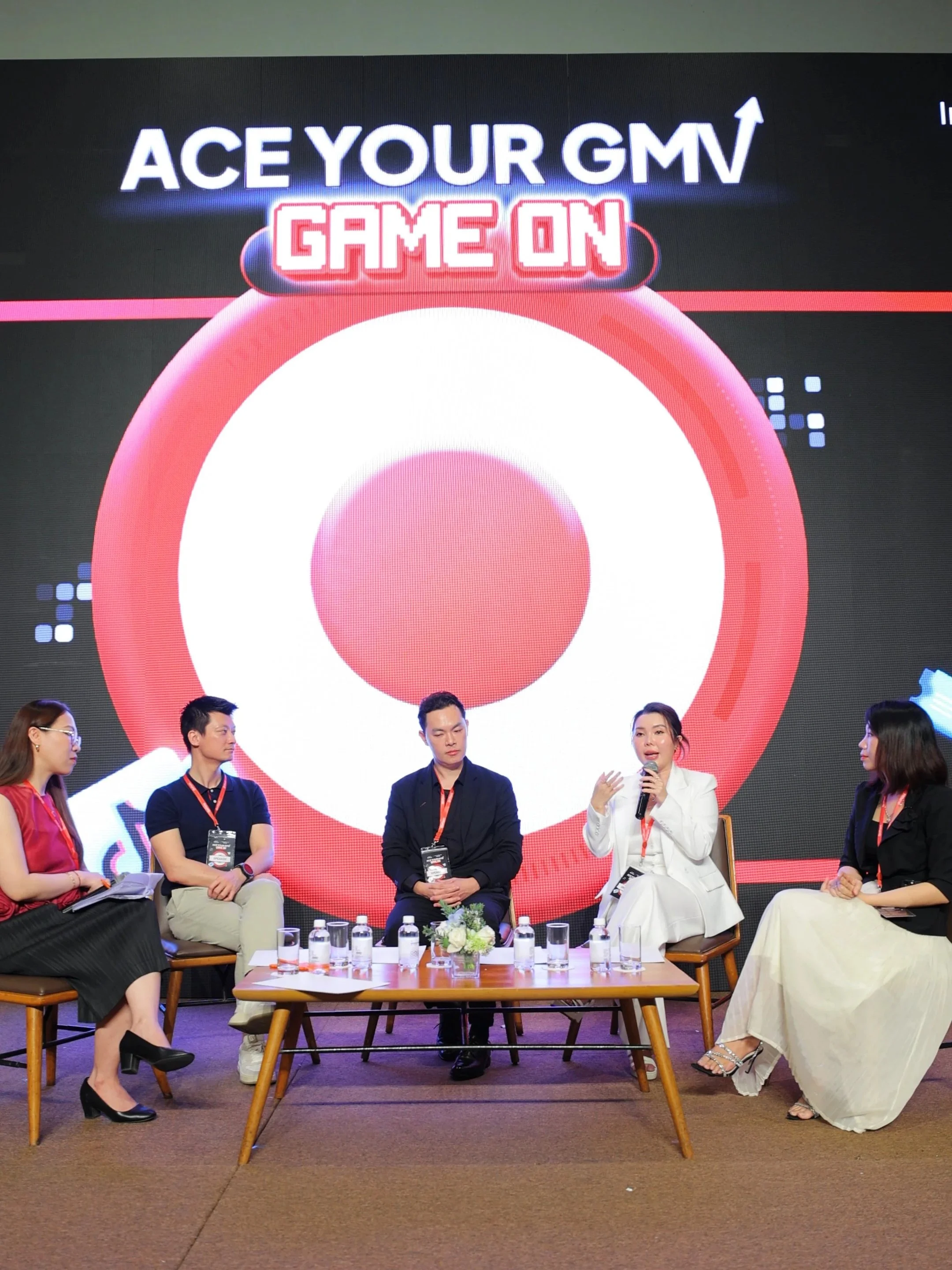 A group of five people sitting on chairs in front of a large screen with the text 'Ace Your GM' and 'Game On'. One woman is speaking into a microphone, and there are water bottles and a flower arrangement on the table in front of them.