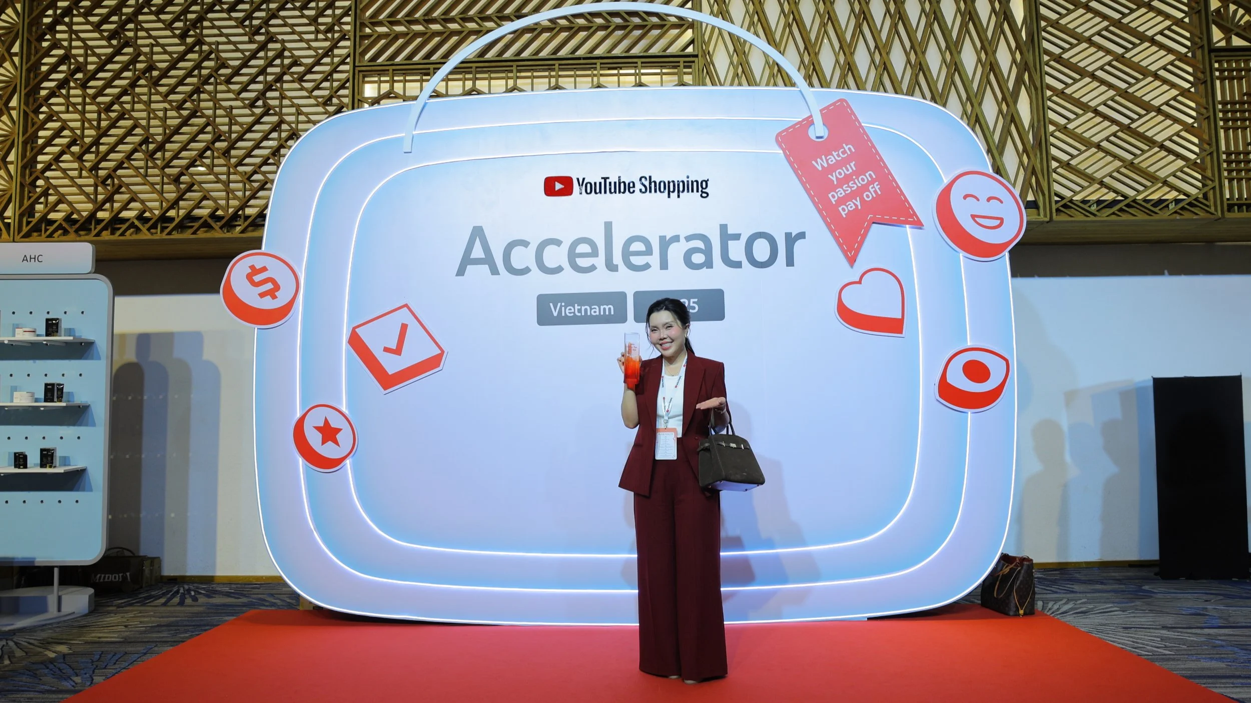 A woman in a maroon suit holding a drink and a handbag at the YouTube Shopping Accelerator event in Vietnam, standing in front of a large illuminated display with the event name and various icons and slogans.