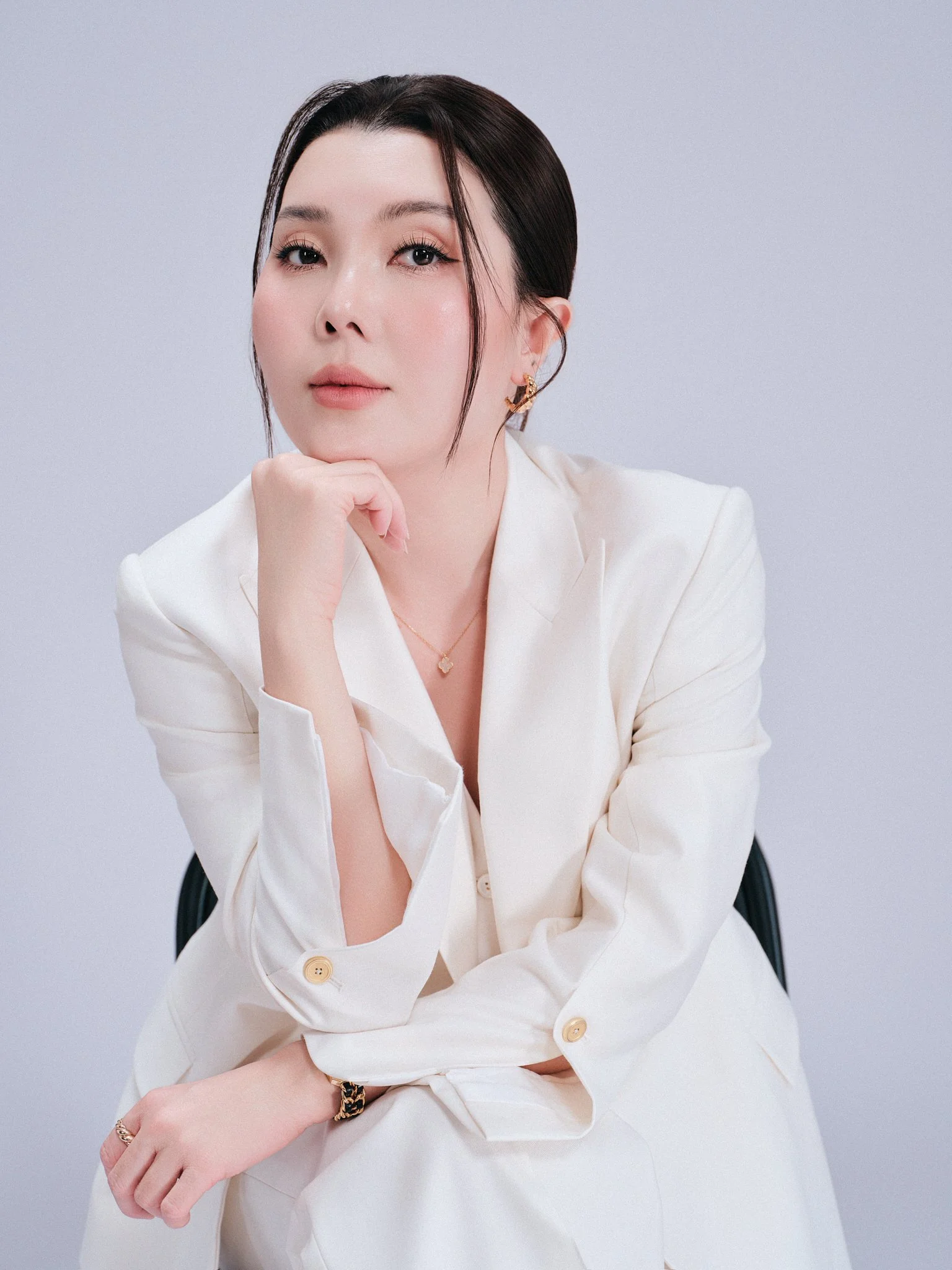 A woman with dark hair styled back, wearing a white blazer and jewelry, sitting and resting her chin on her hand, looking at the camera against a plain light-colored background.