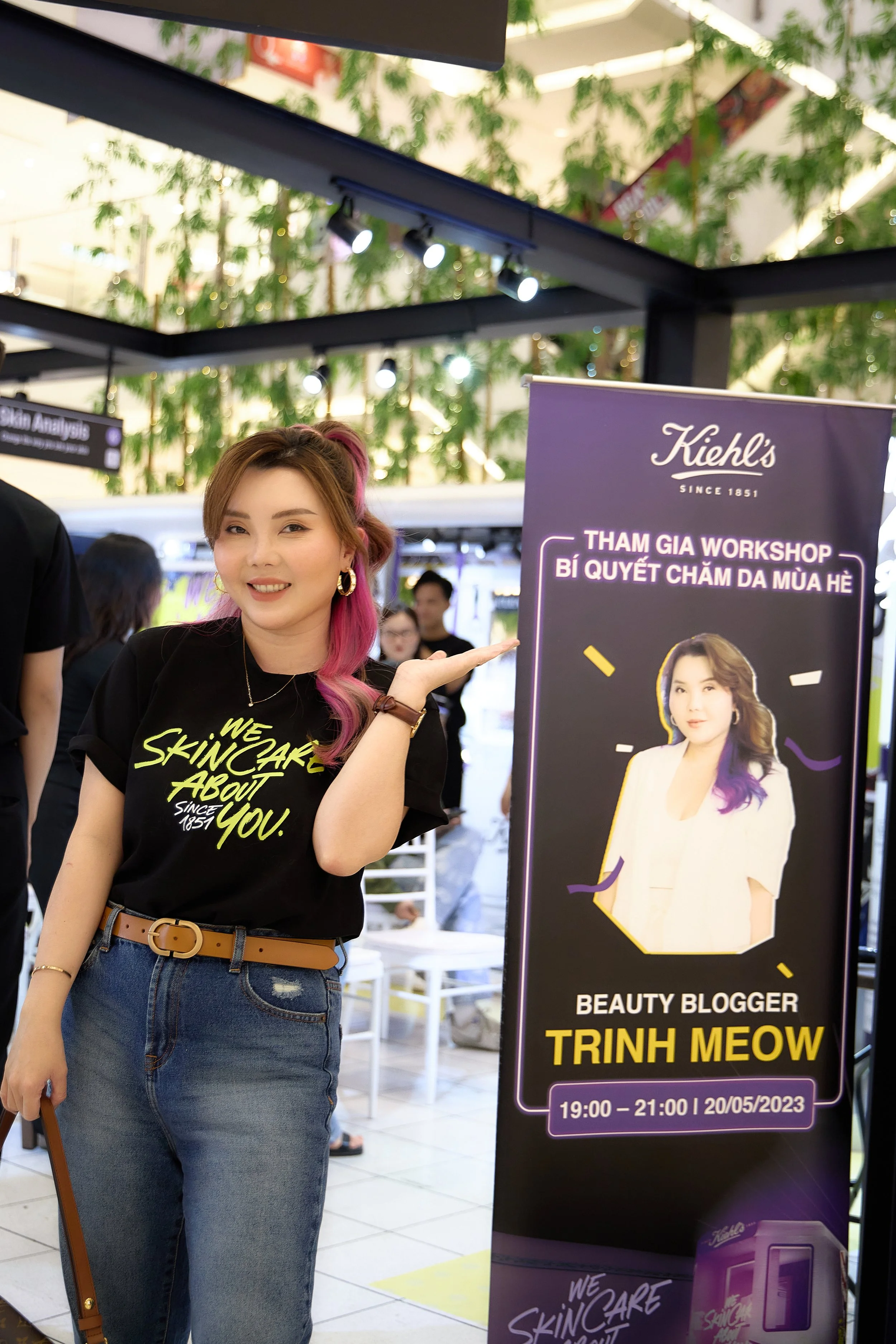 A woman with pink and brown hair in a black T-shirt and jeans standing next to a Kiehl's promotional banner at an indoor event.
