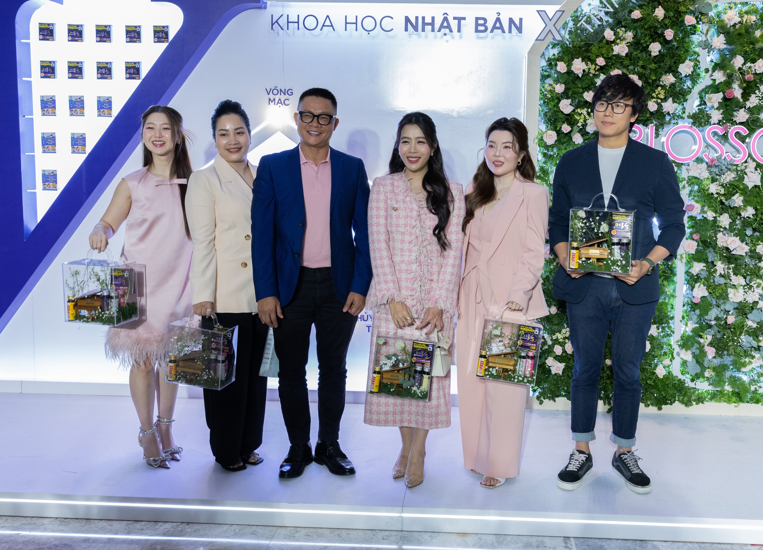 A group of seven people, five women and two men, standing together at an event, holding gift baskets containing beauty or health products. They are dressed in formal or semi-formal attire, smiling, with a decorative green floral backdrop and a white wall with blue accents behind them.