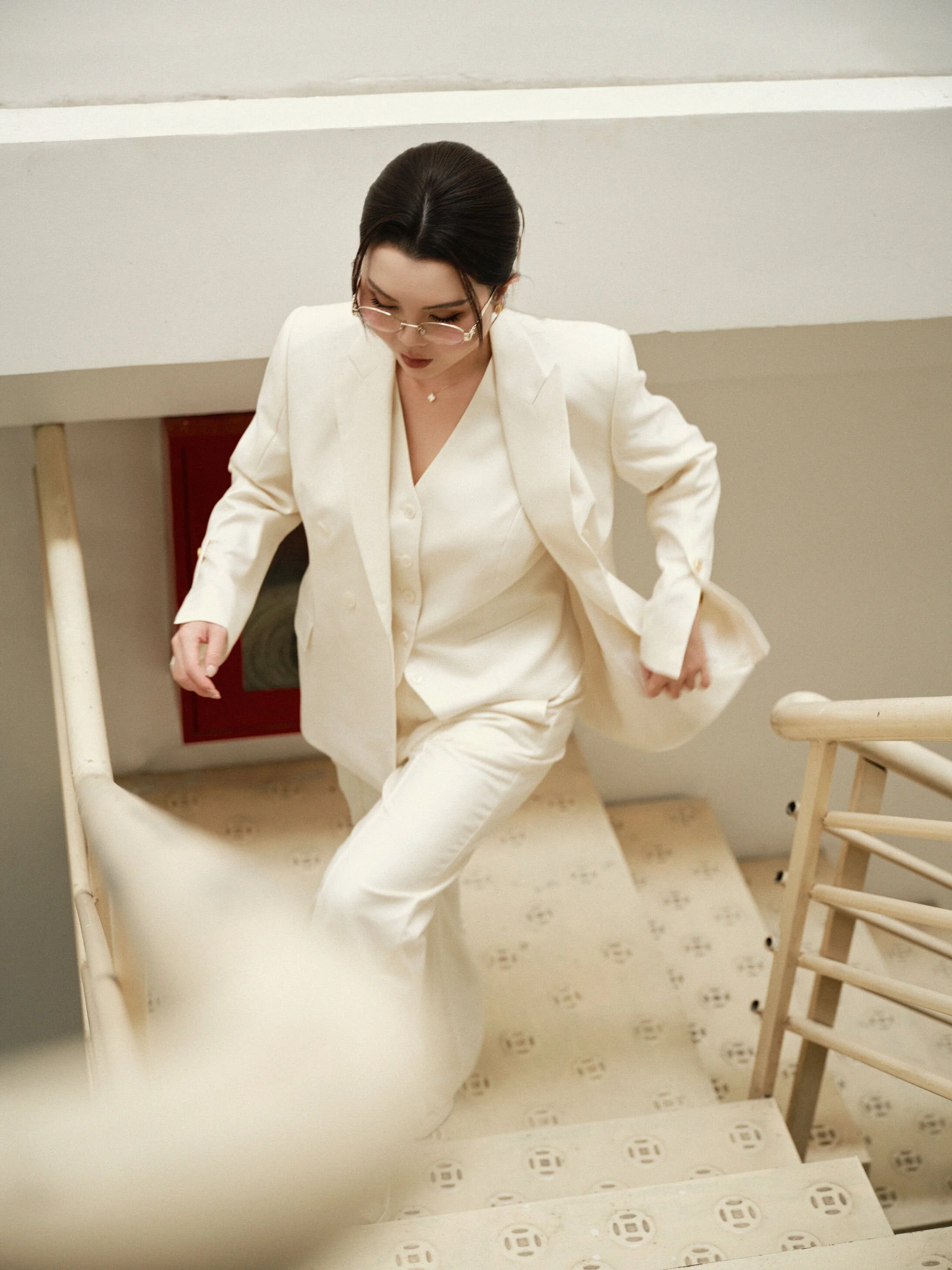 Woman with dark hair, wearing glasses and an all-white suit, climbing up a staircase.