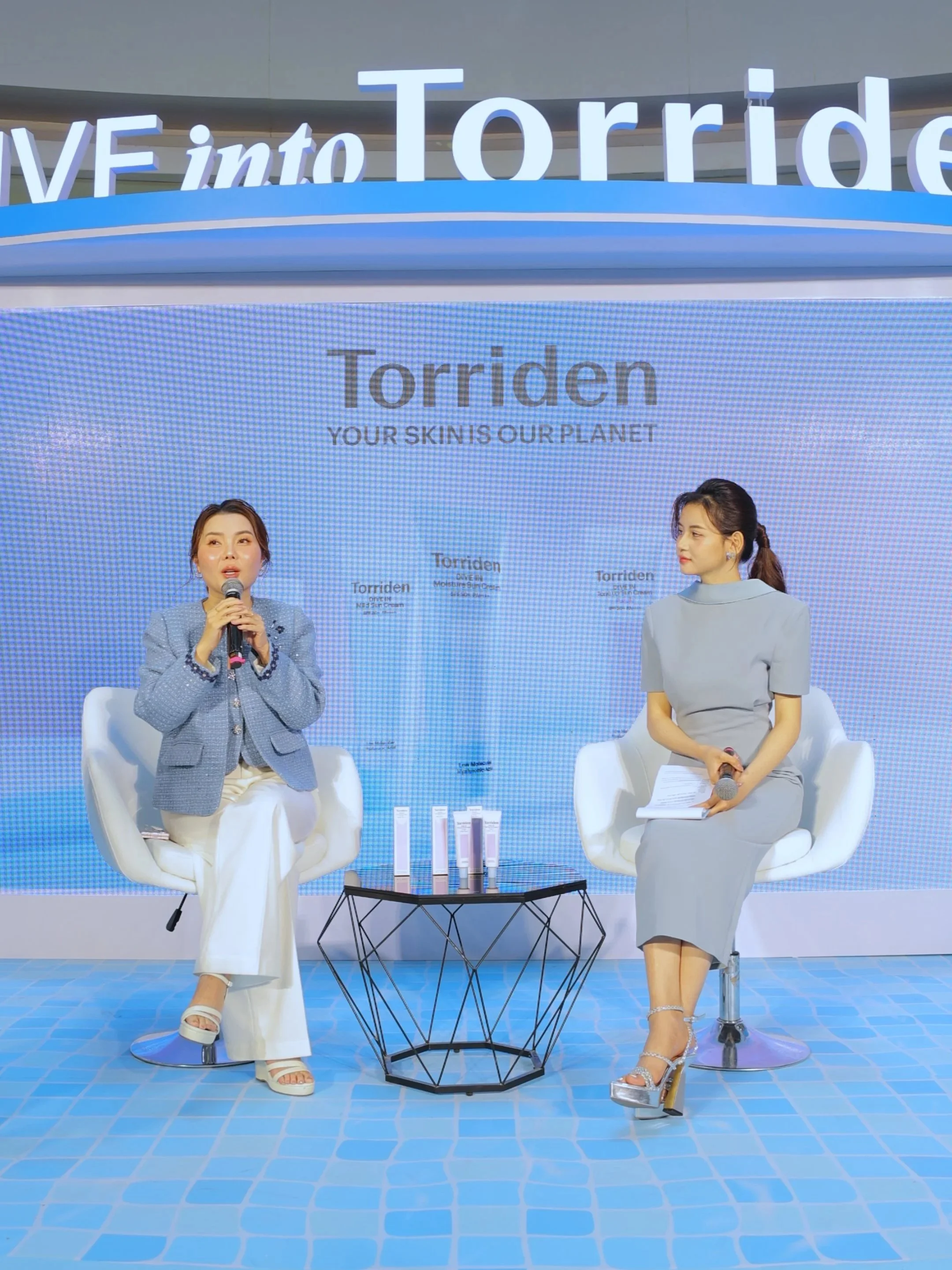 Two women sitting on white chairs in front of a backdrop that says 'Torriden' and 'Your Skin Is Our Planet'. They appear to be at a promotional event or interview, with a small table holding skincare products between them. The woman on the left is speaking into a microphone, dressed in a blue blazer and white pants, while the woman on the right is listening, dressed in a light gray dress.