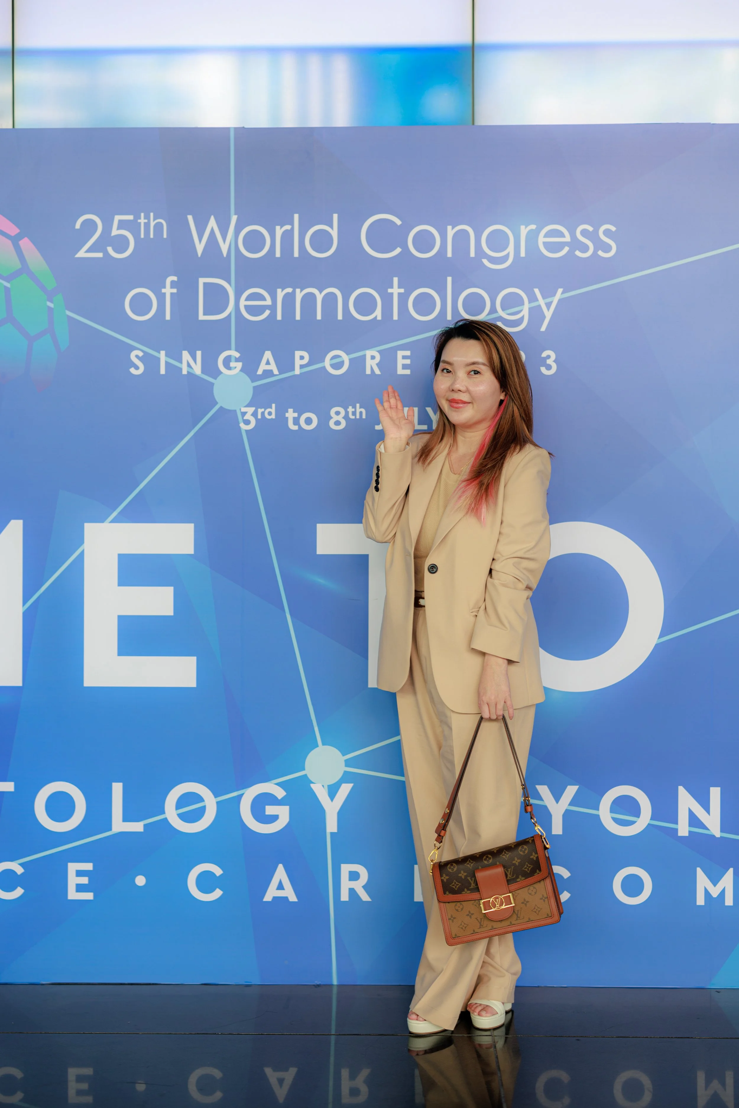 A woman standing in front of a large blue banner for the 25th World Congress of Dermatology in Singapore, from July 3 to 8, 2023. She is wearing a beige suit, holding a Louis Vuitton handbag, and smiling at the camera.