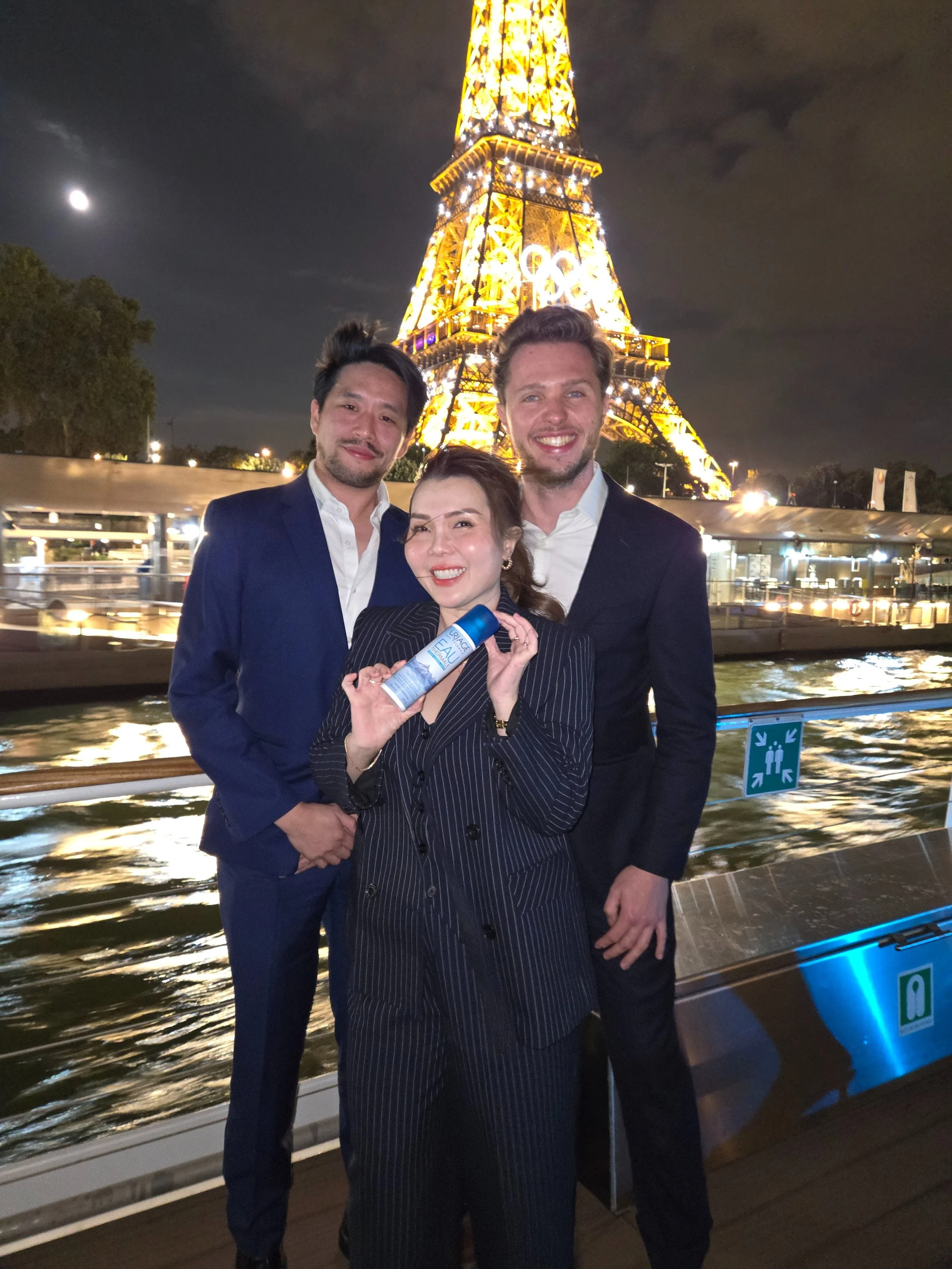 Three people smiling at the camera on a boat near the Eiffel Tower at night, with the city lights and river visible.