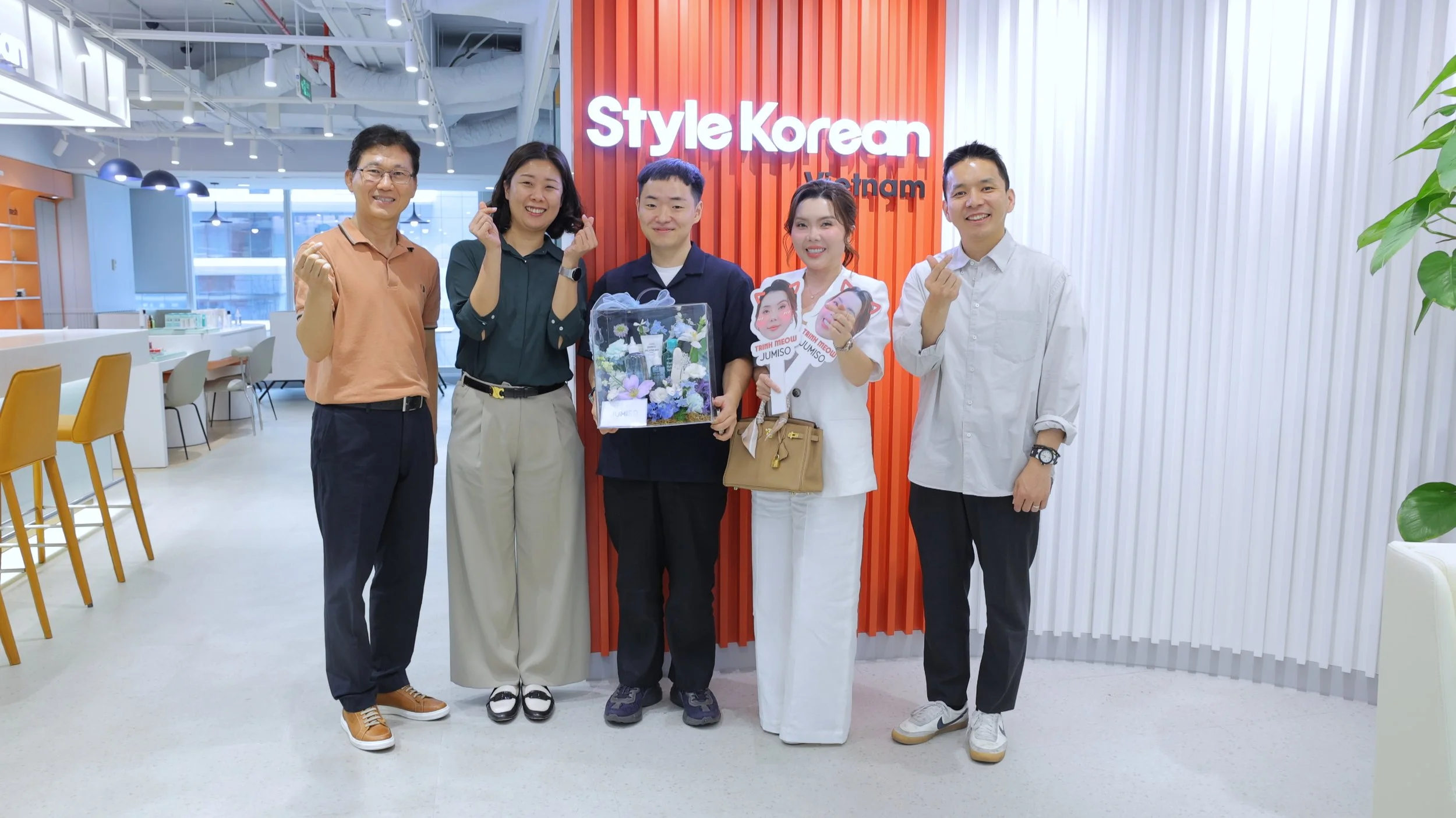 Group of five people standing inside an office, smiling, with a red wall behind them that has the sign 'Style Korean Vietnam'. One woman holds a gift box decorated with flowers, and another holds photo props.