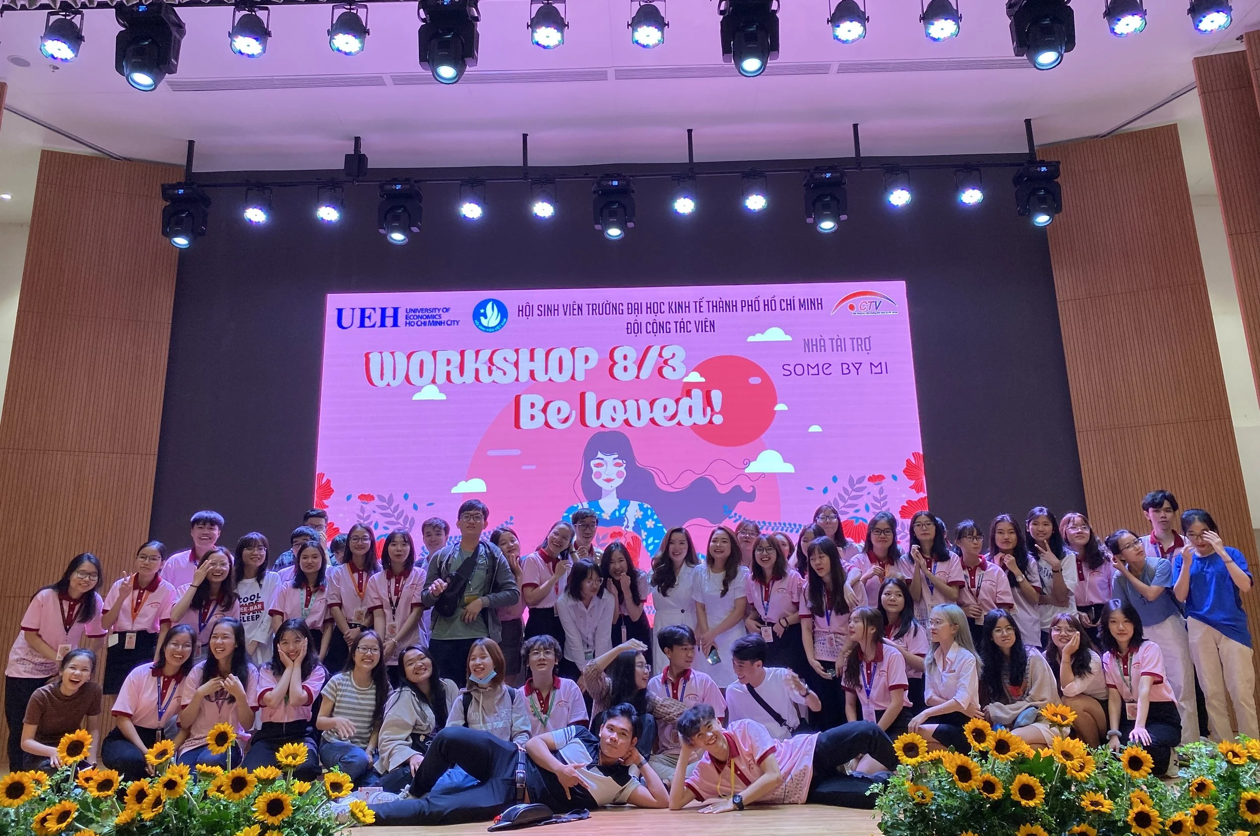 A group of students on a stage in front of a large screen with a colorful backdrop. The backdrop features a girl with long hair, clouds, a red sun, and the text 'WORKSHOP 8/3 Be loved!' and other information in Vietnamese. The stage is decorated with sunflower plants at the front.