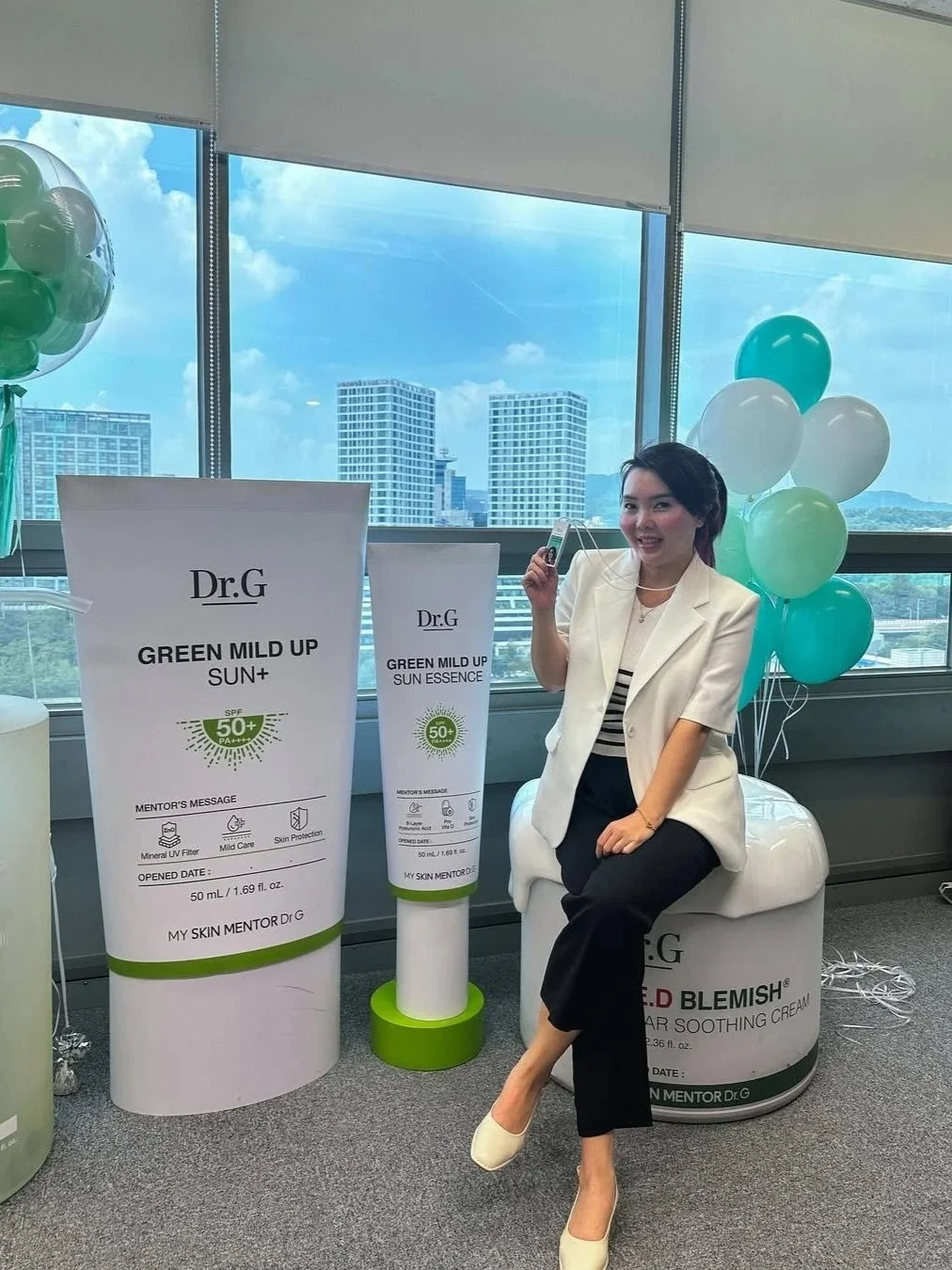 Woman sitting on a white skincare product container at a promotional event with large product displays and balloons in the background.