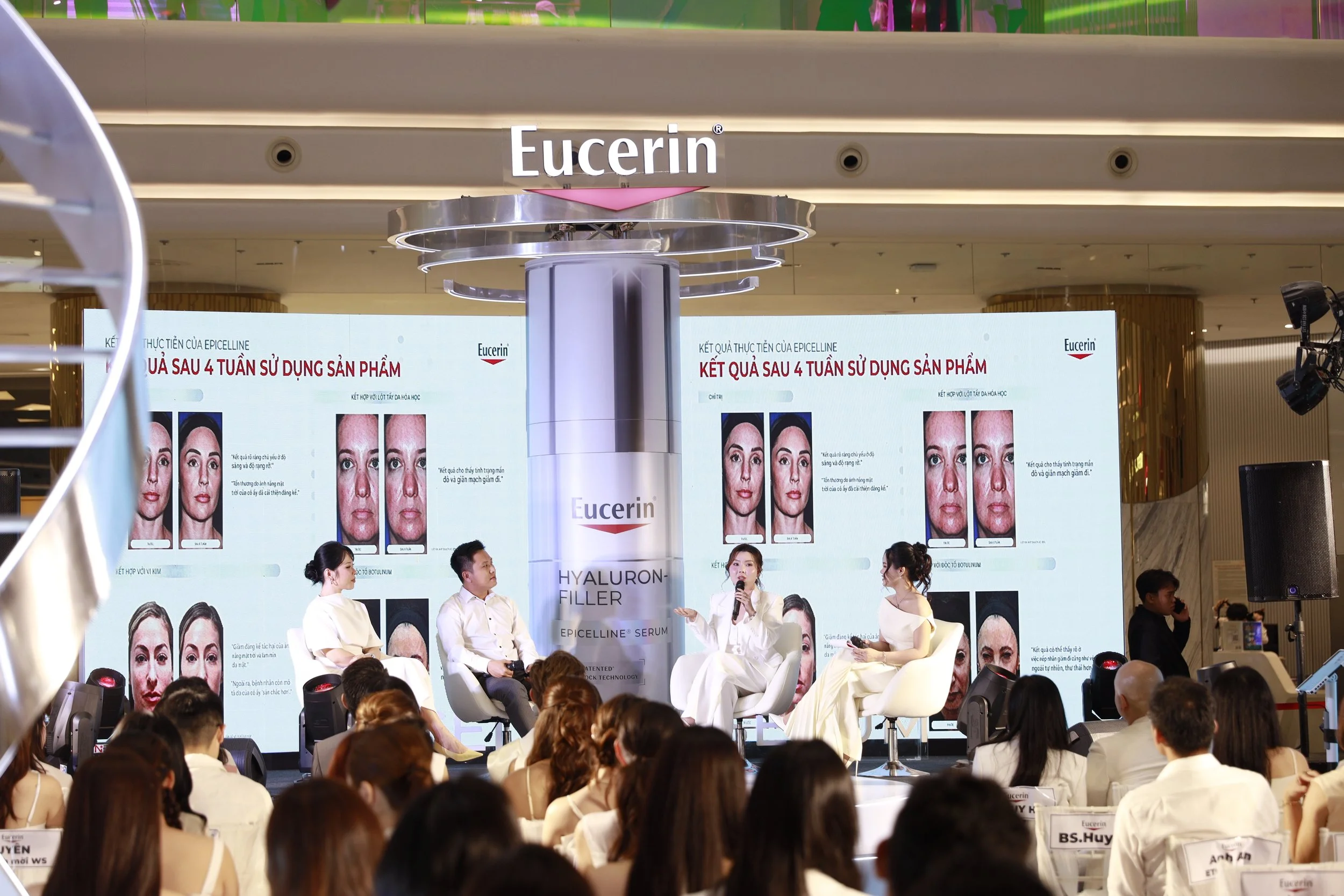 A panel discussion at a conference with four speakers on stage, two women and two men, sitting on white chairs. Behind them is a large screen displaying the Eucerin logo and image comparisons showing the results after 4 weeks of using a skincare product. The audience is seated in front, and there is a large Eucerin sign hanging above the stage.