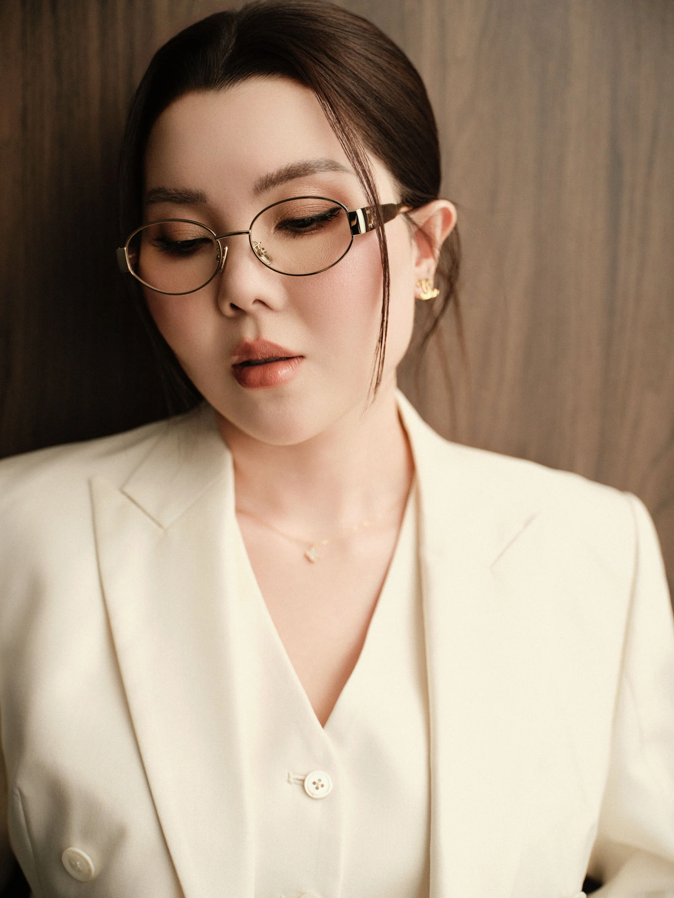 A woman with dark hair, wearing glasses, gold earrings, and a cream-colored blazer, looking down with a neutral expression in front of a wooden background.