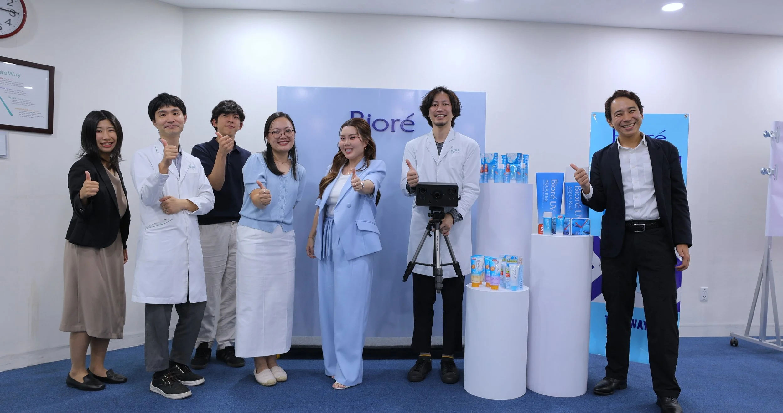 Group of seven people, including men and women, standing indoors, giving thumbs-up, in front of a display with products and a sign that reads 'Biøre', some wearing lab coats and business attire, smiling at the camera.