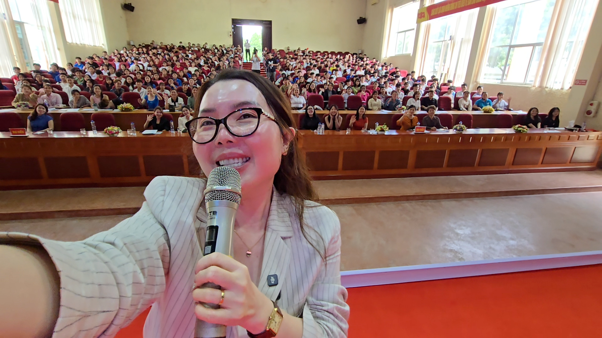 A woman taking a selfie on stage with a microphone in front of a large audience in an auditorium, several rows of people smiling and waving.