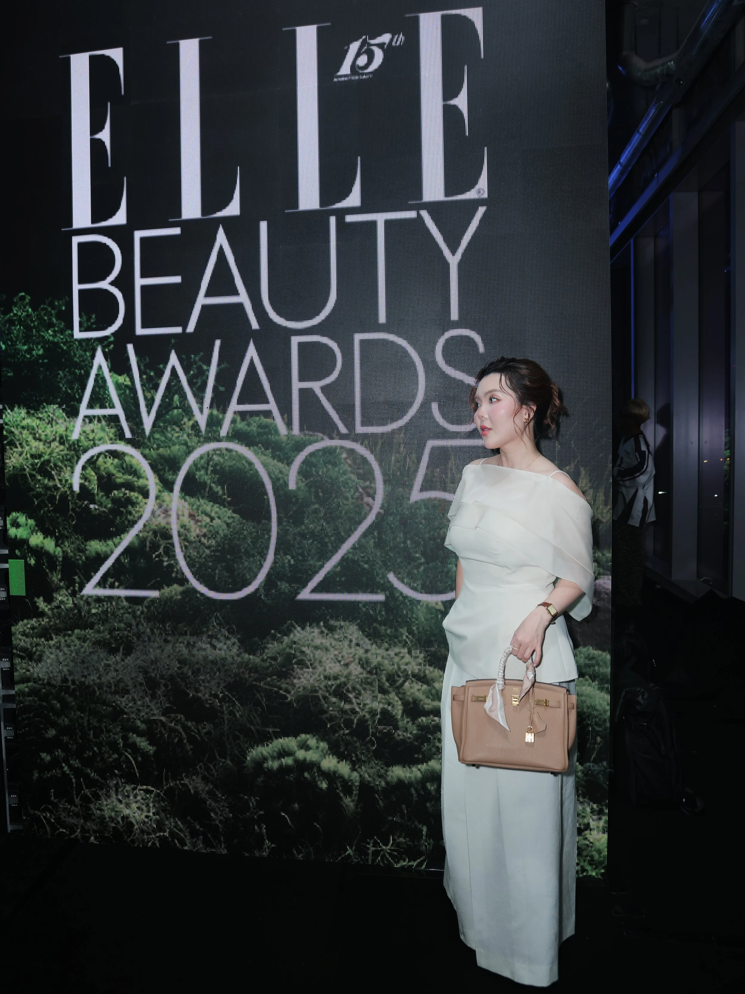 A woman in an off-the-shoulder white dress holding a beige handbag, standing in front of a large sign that reads 'ELLE Beauty Awards 2023', during an event.