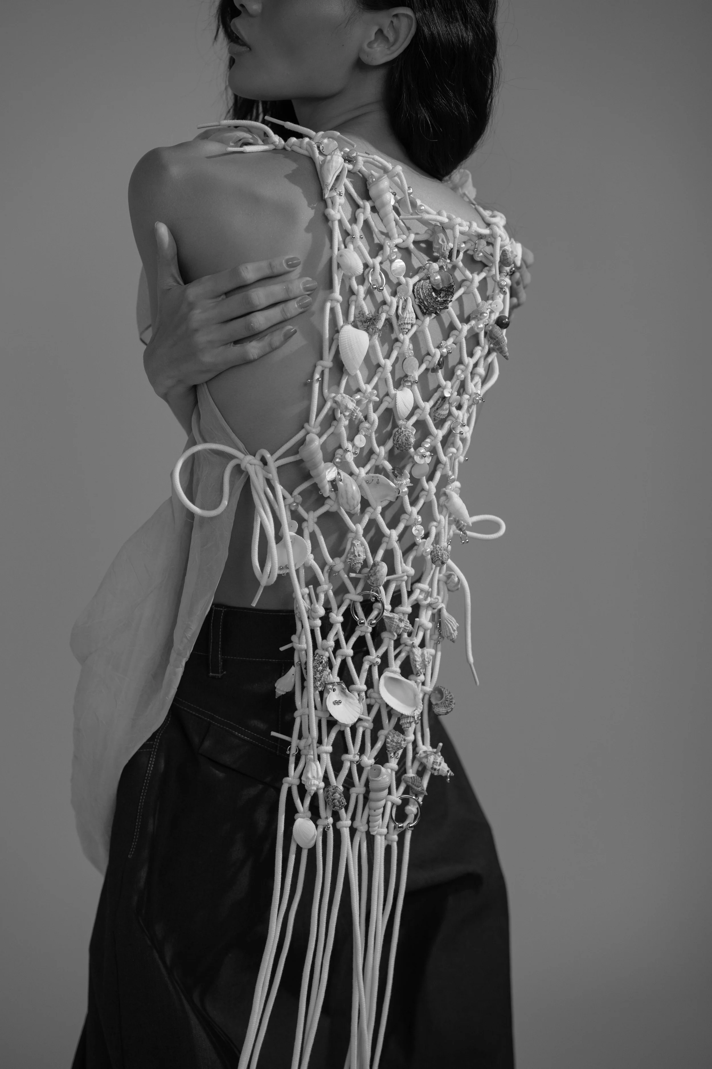 Black and white photo of a woman wearing an intricate netted garment made of ropes and adorned with seashells, beads, and small trinkets.