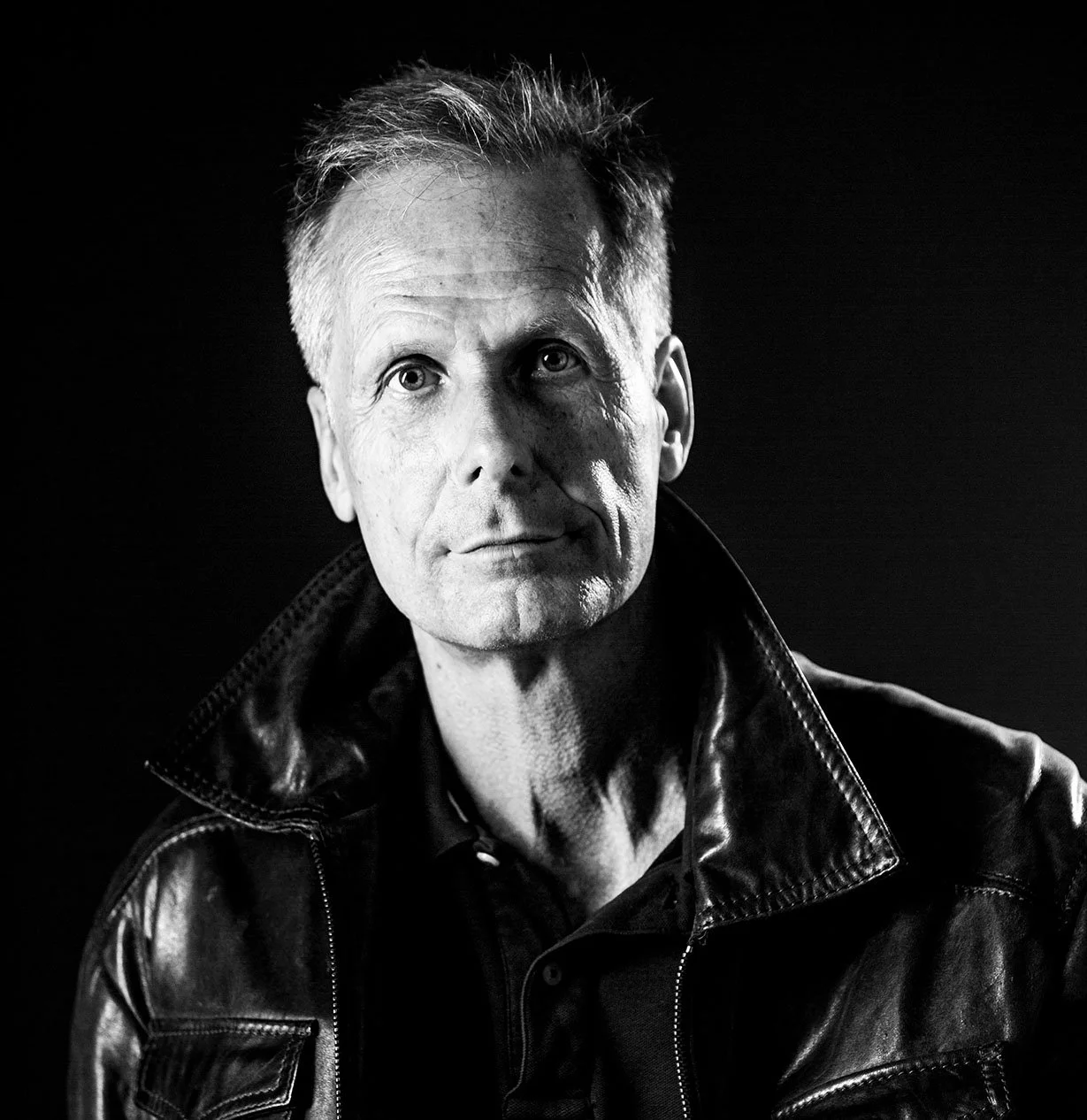 Schwarz-Weiß-Porträt eines Mannes mit kurzer, glatter, grauer Frisur, der eine Lederjacke trägt, gegen schwarzen Hintergrund.