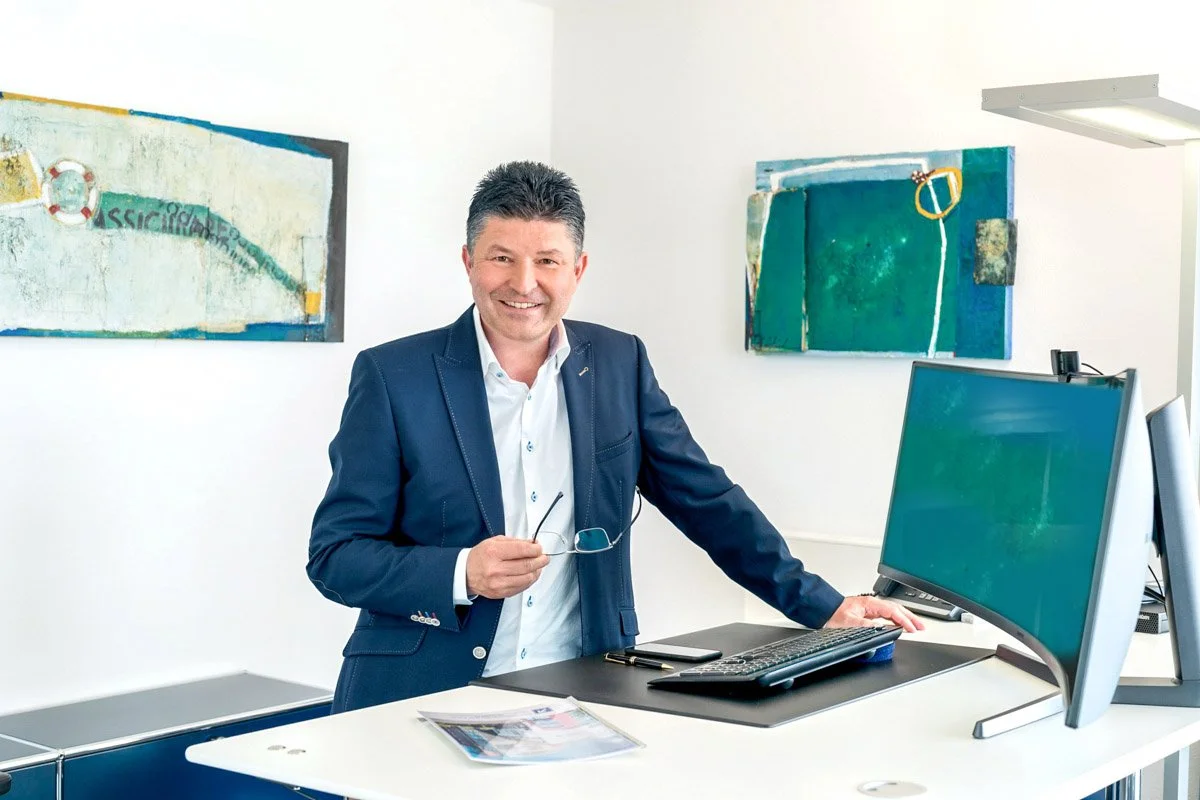 Ein Mann in einem Business-Anzug steht an einem Schreibtisch in einem modernen Büro, lächelt und hält eine Brille in der Hand. Im Hintergrund sind zwei bunte, abstrakte Gemälde an der Wand zu sehen. Auf dem Schreibtisch befinden sich ein Computer, eine Tastatur, eine Maus, ein Smartphone und einige Unterlagen.