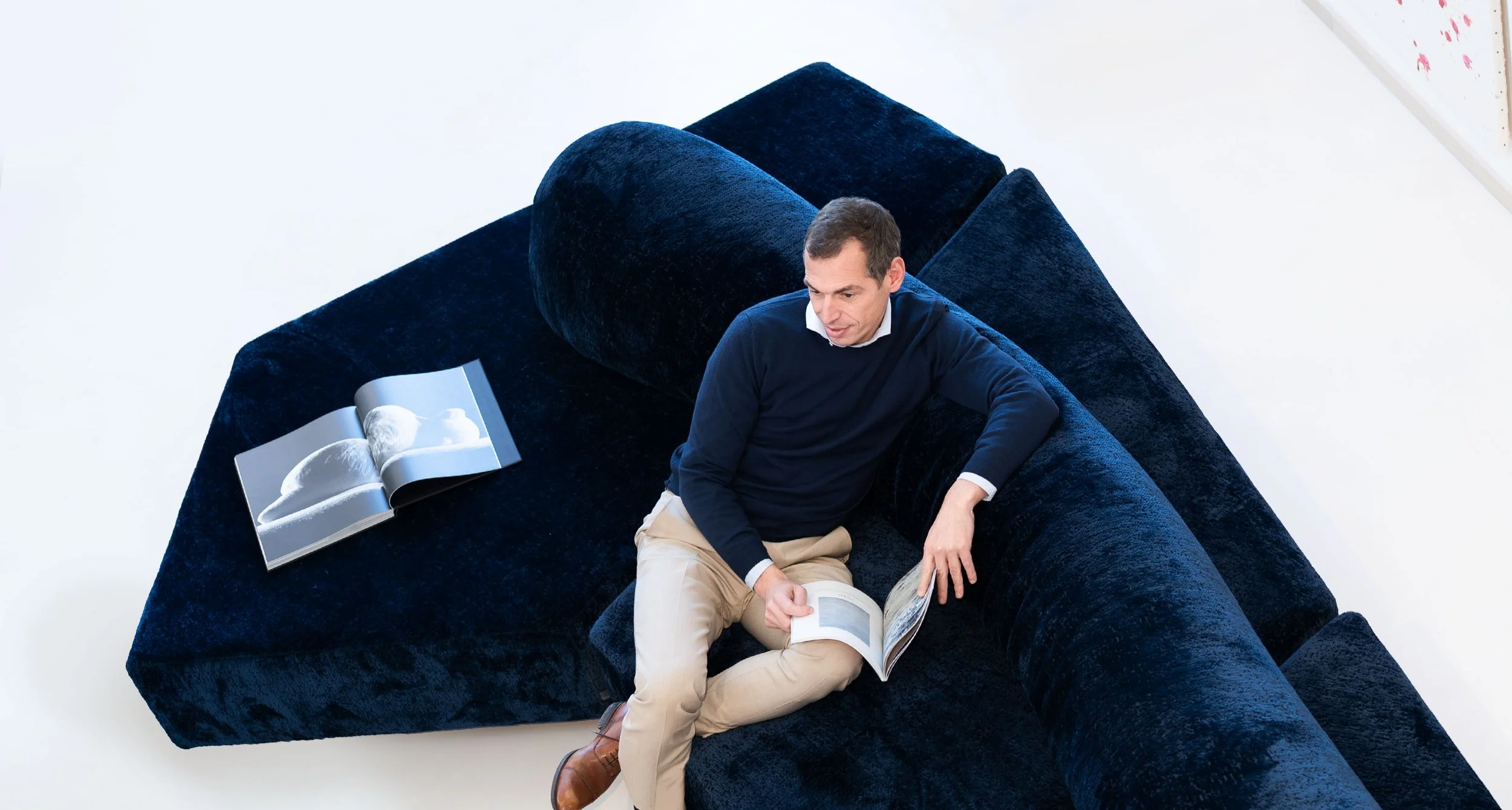 Mann sitzt auf einer dunkelblauen Couch und liest ein Buch, ein weiteres Buch liegt neben ihm auf der Couch.