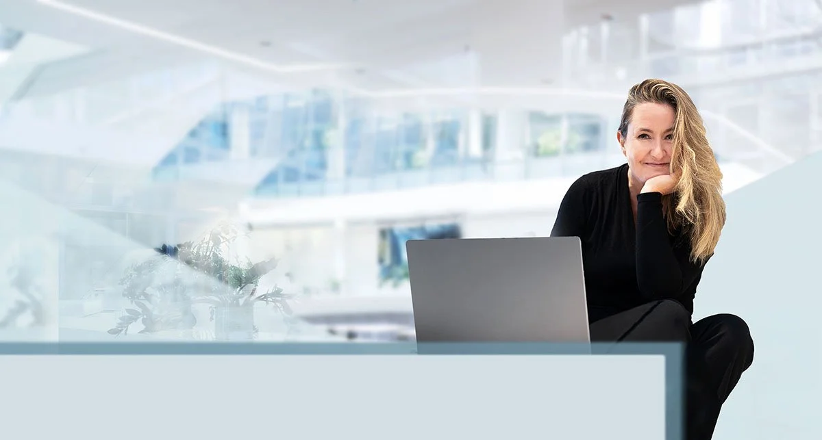 Eine Frau sitzt mit einem Laptop in einem modernen, hellen Büro. Sie lächelt leicht und hat lockiges Haar. Im Hintergrund sind große Fenster und eine moderne Architektur zu sehen.