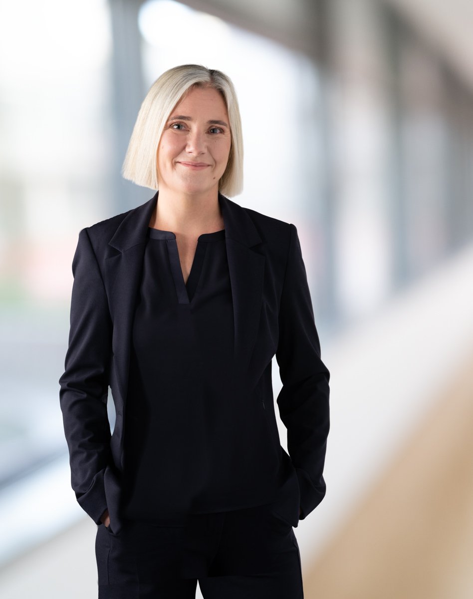 Eine junge Frau mit blonder Bobfrisur und dunkler Kleidung steht in einem modernen, hell erleuchteten Raum. Sie lächelt leicht und hat die Hände in den Taschen.