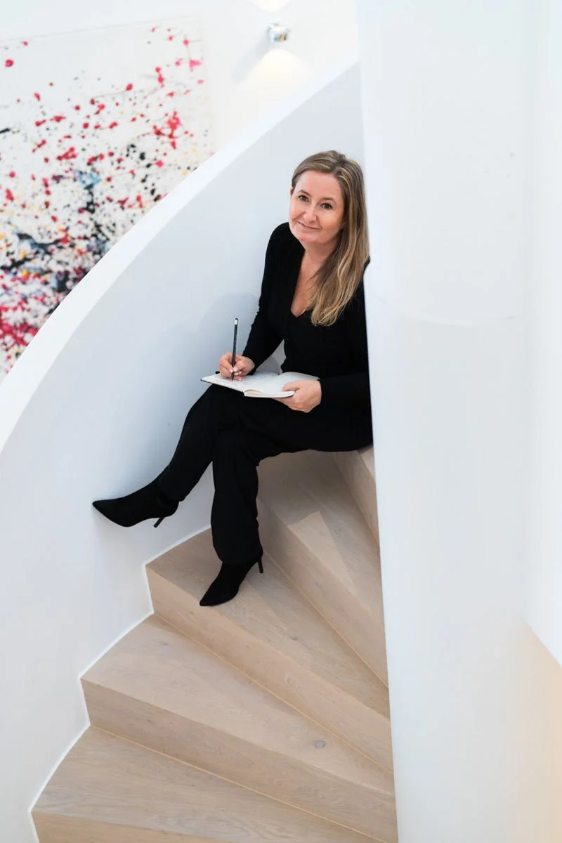 Eine Frau in schwarzer Kleidung sitzt auf einer Treppe im Spiraldesign, hält ein Notizbuch und schreibt mit einem Stift. Sie blickt nach oben, im Hintergrund ist eine bunte abstrakte Wandkunst zu sehen.