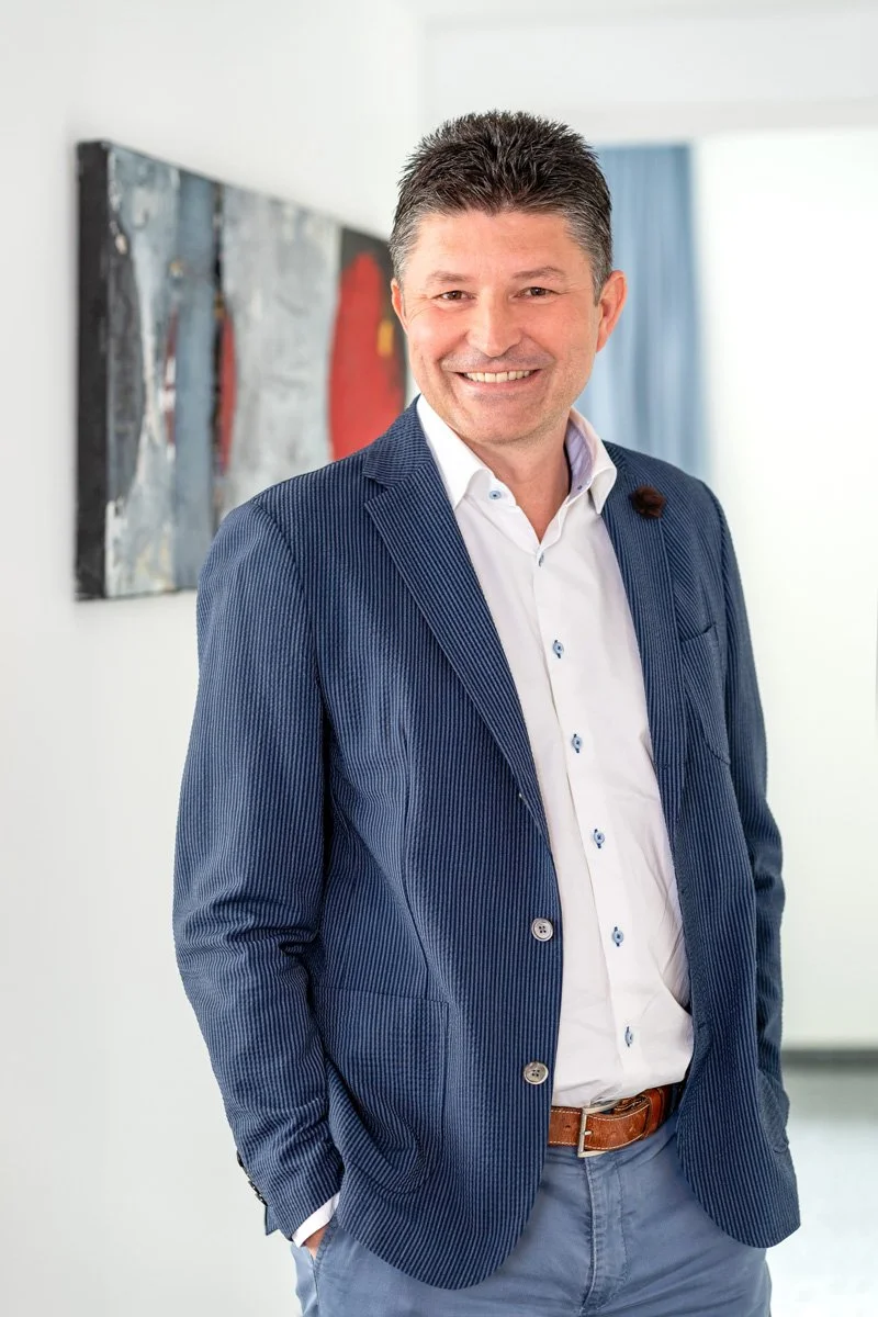 Ein Mann in einem blauen Anzug mit weißem Hemd, lächelnd, steht vor einer weißen Wand mit abstrakter Kunst an der Seite.