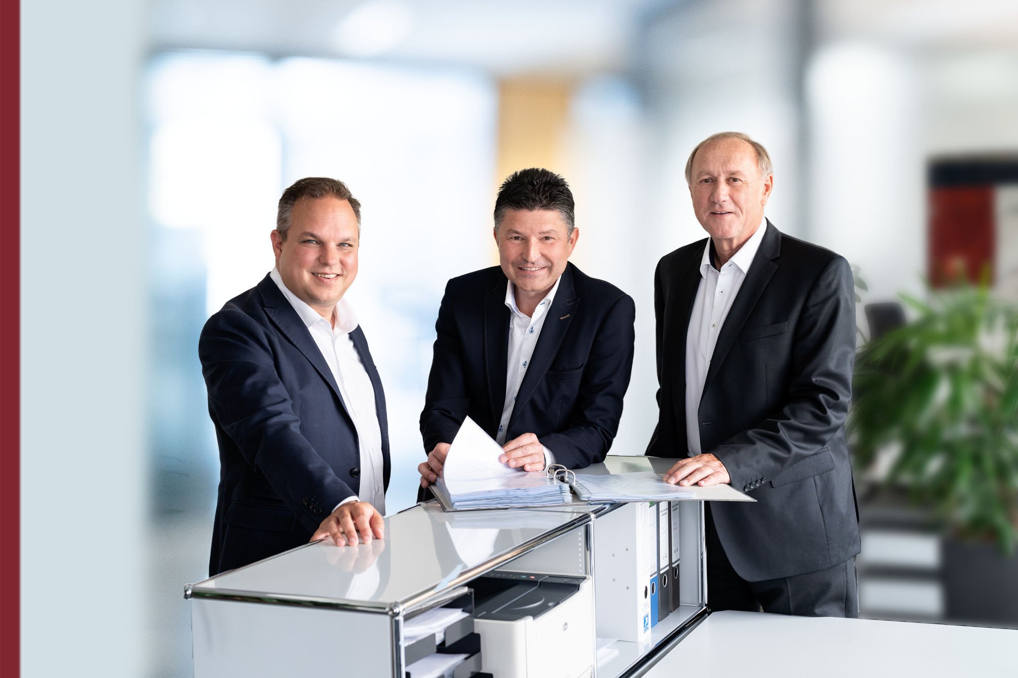 Drei Männer in Anzügen stehen um einen Schreibtisch mit Aktenordnern, in einem modernen Büro.