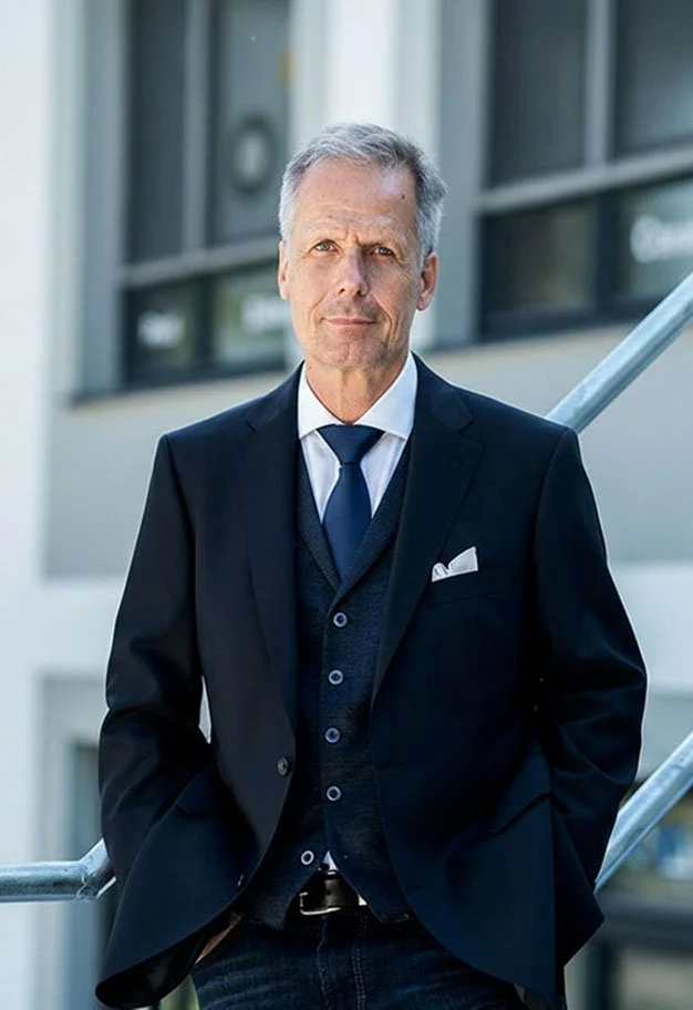 Ein älterer Mann in einem dunklen Anzug steht vor einem modernen Gebäude, mit seinem rechten Arm an einem Geländer. Er blickt ernst in die Kamera, trägt ein weißes Hemd, eine schwarze Krawatte und eine dunkelblaue Weste.