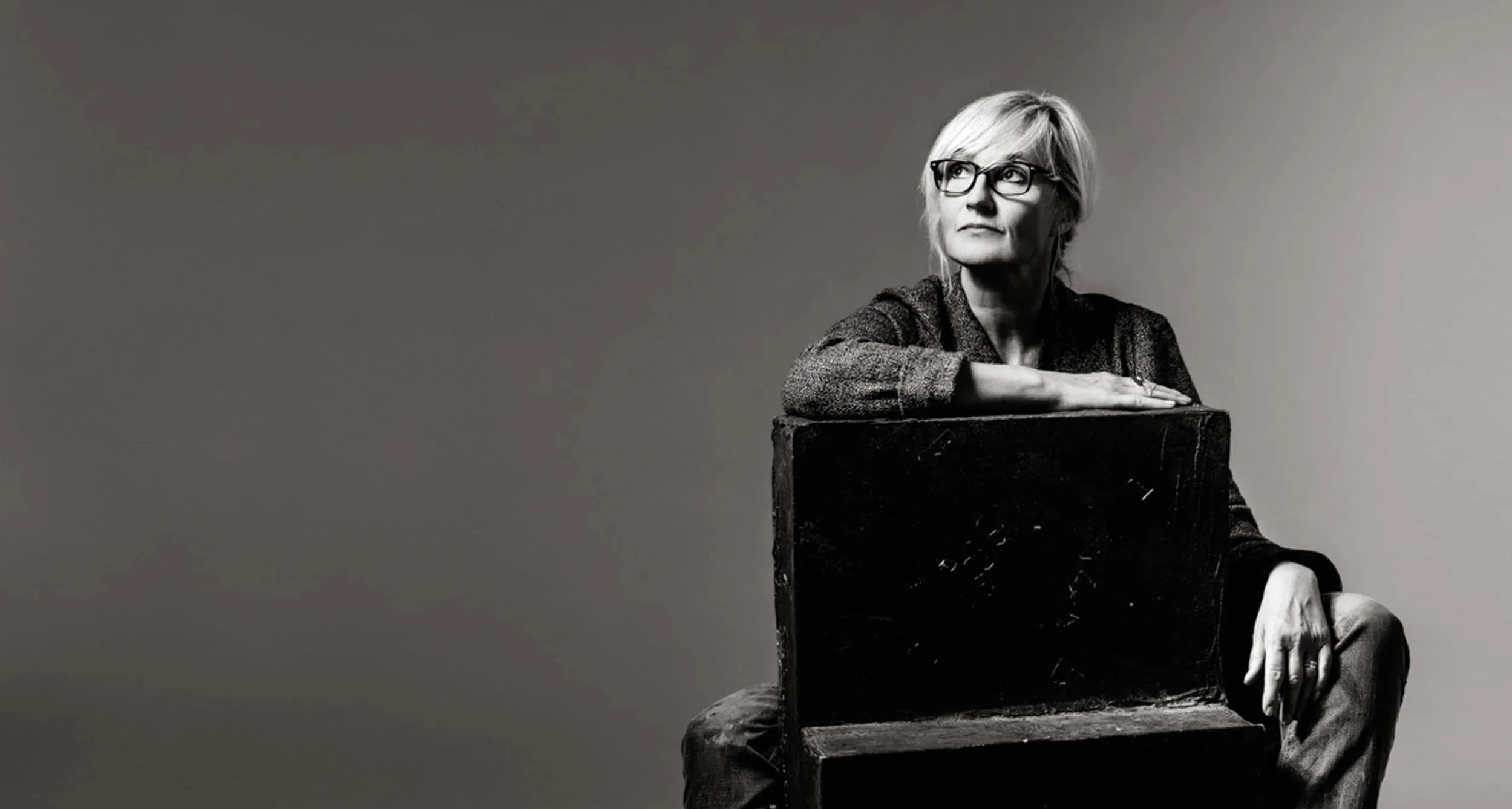 Schwarz-weiß Porträt einer Frau mit Kurzhaarfrisur und Brille, die auf einem Stuhl sitzt, den Arm auf die Rückenlehne gelegt, nachdenklich blickend nach rechts.