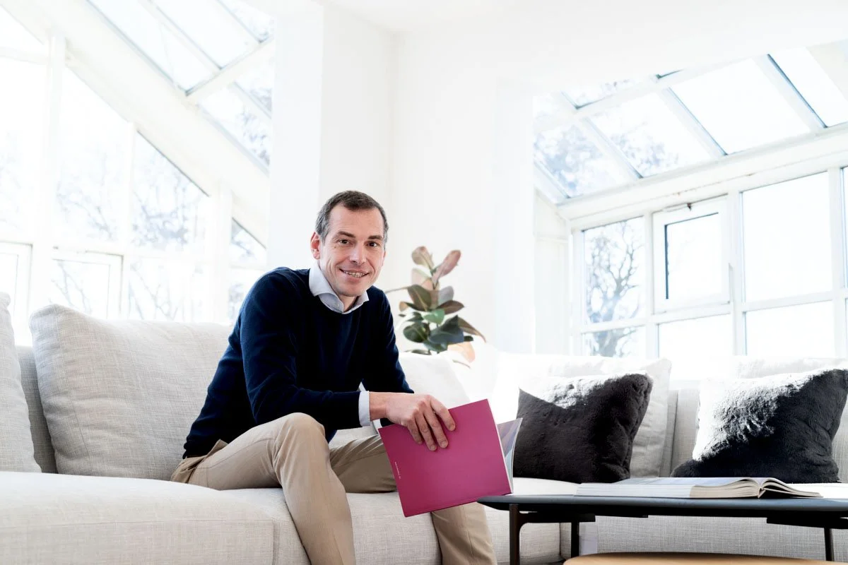 Ein Mann sitzt auf einem weißen Sofa in einem hellen Raum mit großen Fenstern und liest ein pinkfarbenes Buch.